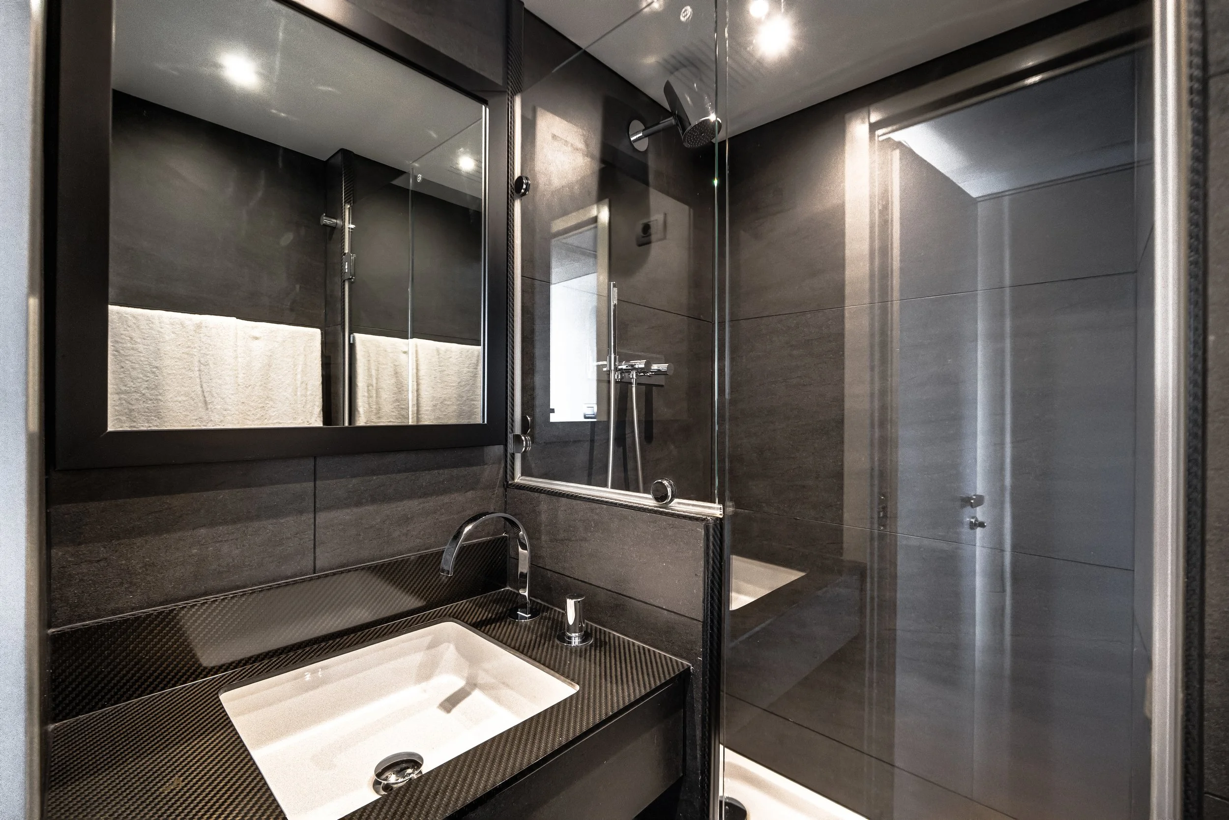 A modern bathroom with a glass-enclosed shower, dark gray tiles, a handheld showerhead, a small white sink, and a mirror. LYFE AT SEA