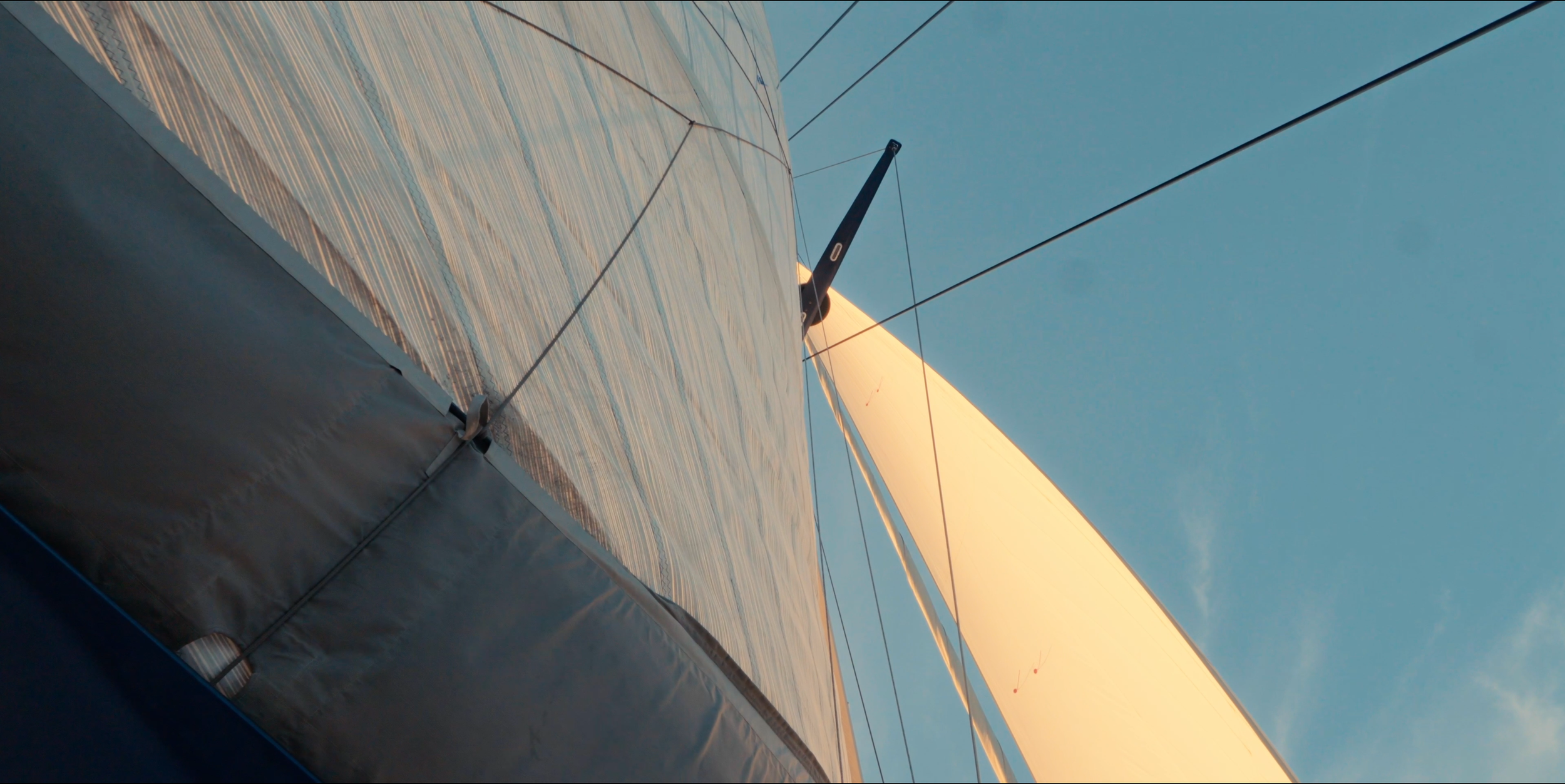 Close-up view of a sailboat's boom, sails, and rigging against a clear blue sky, taken from below the boat. LYFE AT SEA
