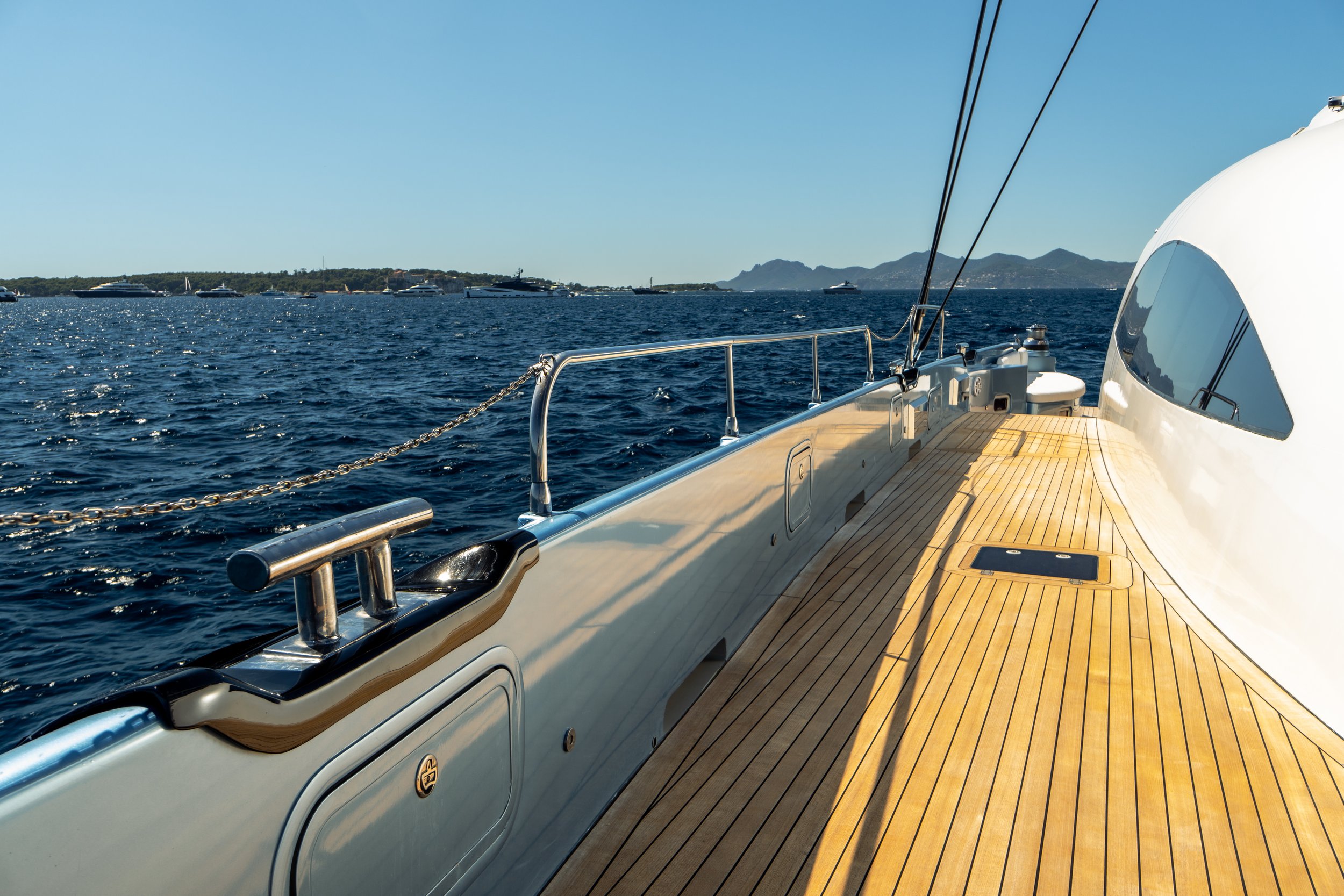 The image shows the deck of a yacht with wood flooring, metal railings, and a smooth white exterior. In the background, there is a large body of water with several other yachts and boats, and distant mountains under a clear blue sky. LYFE AT SEA