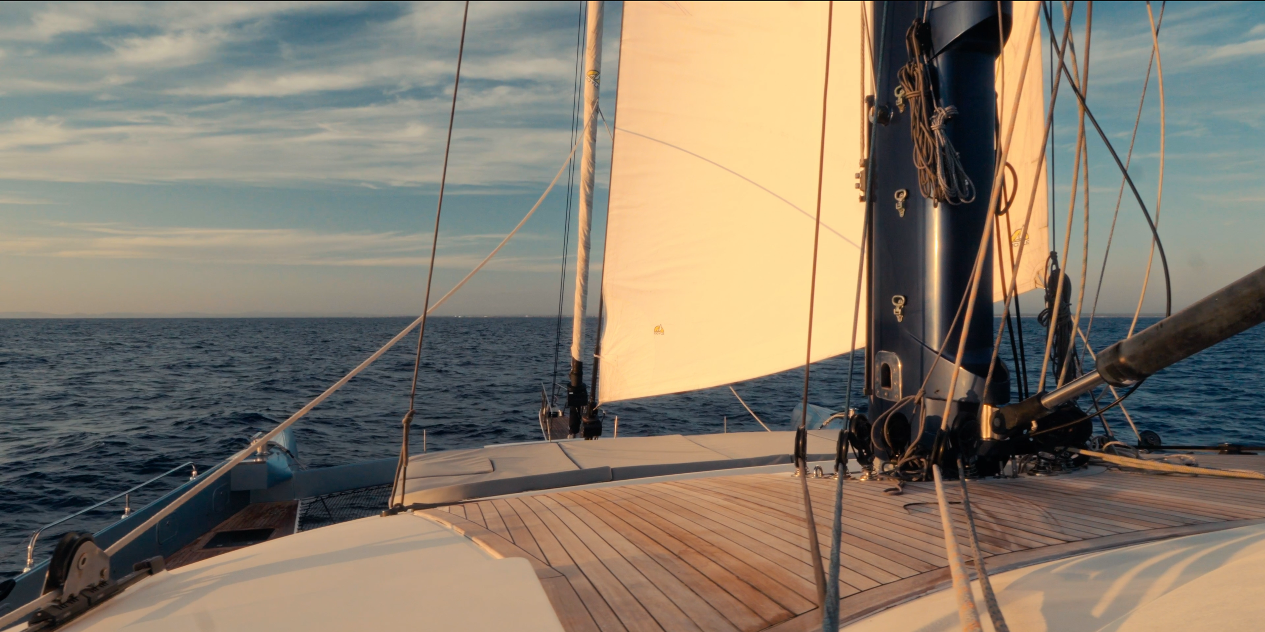 View from the deck of a sailboat on the open ocean with a large white sail, rigging, and a blue hull during sunset. LYFE AT SEA