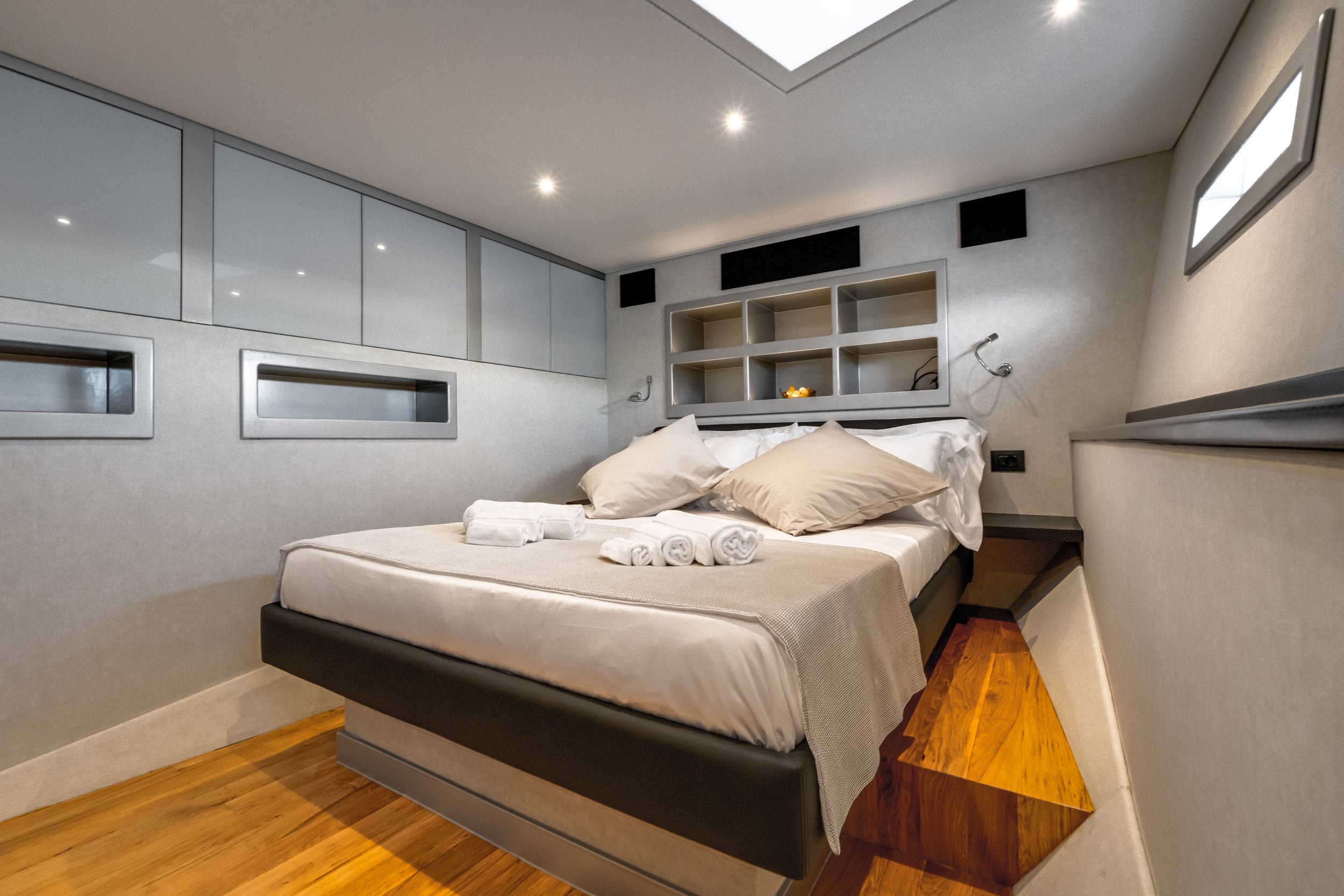 Modern bedroom with a low bed, beige and white bedding, and a small pile of rolled towels. The room has built-in storage, a wooden side table, and overhead lighting.