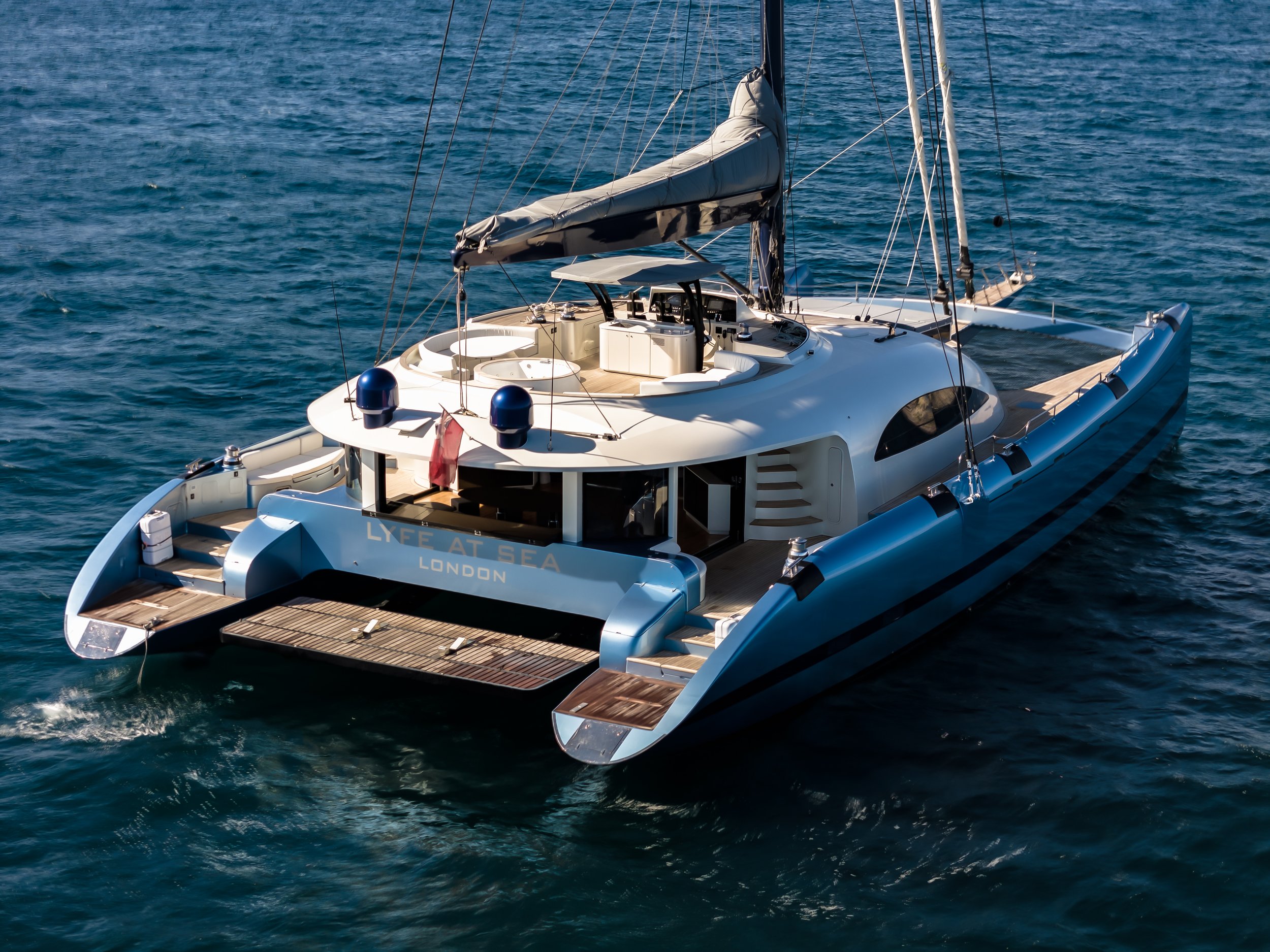 A luxurious white yacht sailing on blue ocean water with visible decks, steering area, and sails. LYFE AT SEA