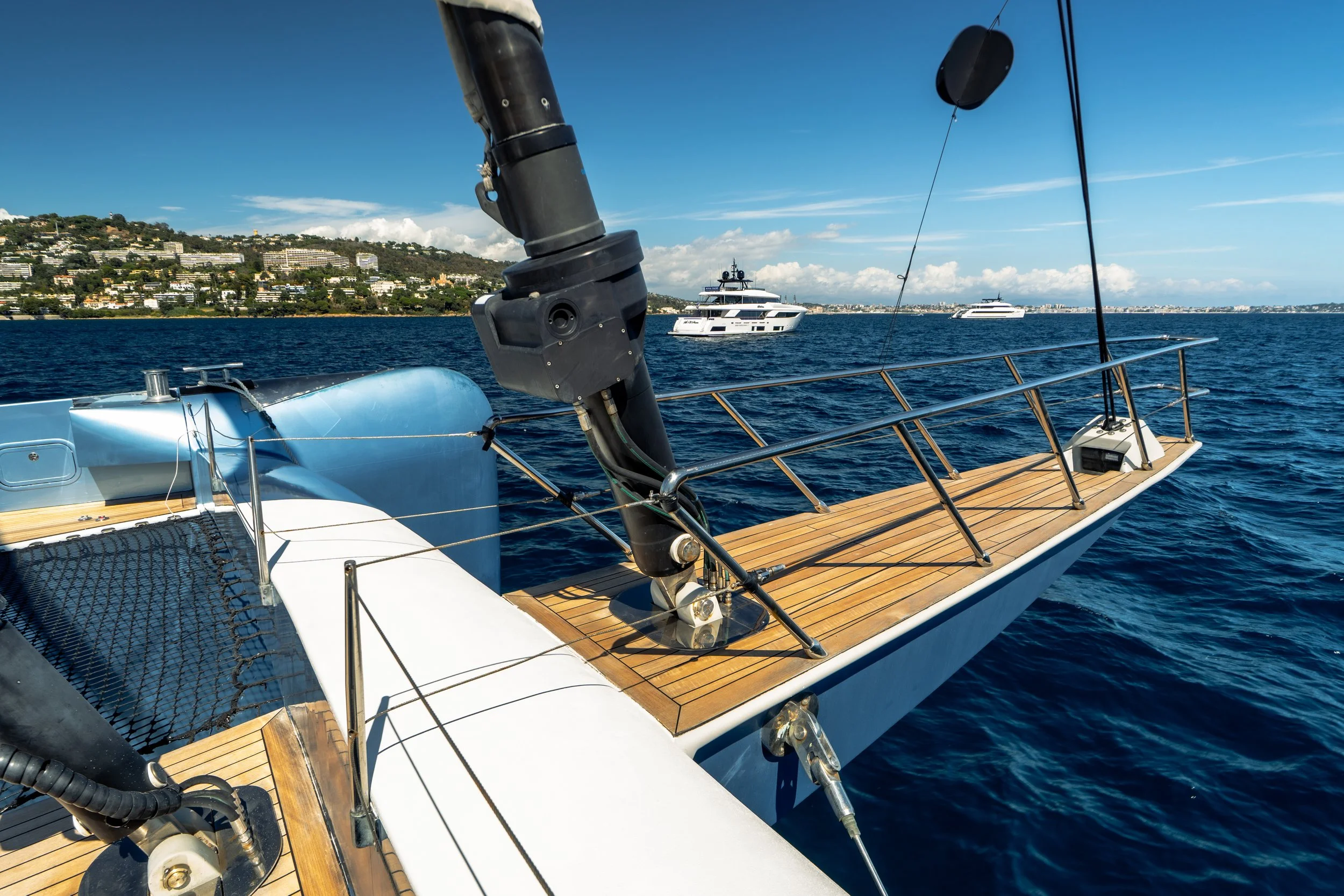 View from a sailboat's deck showing a fishing pole, a yacht in the water, with a hill and buildings in the background, under a blue sky with some clouds. LYFE AT SEA