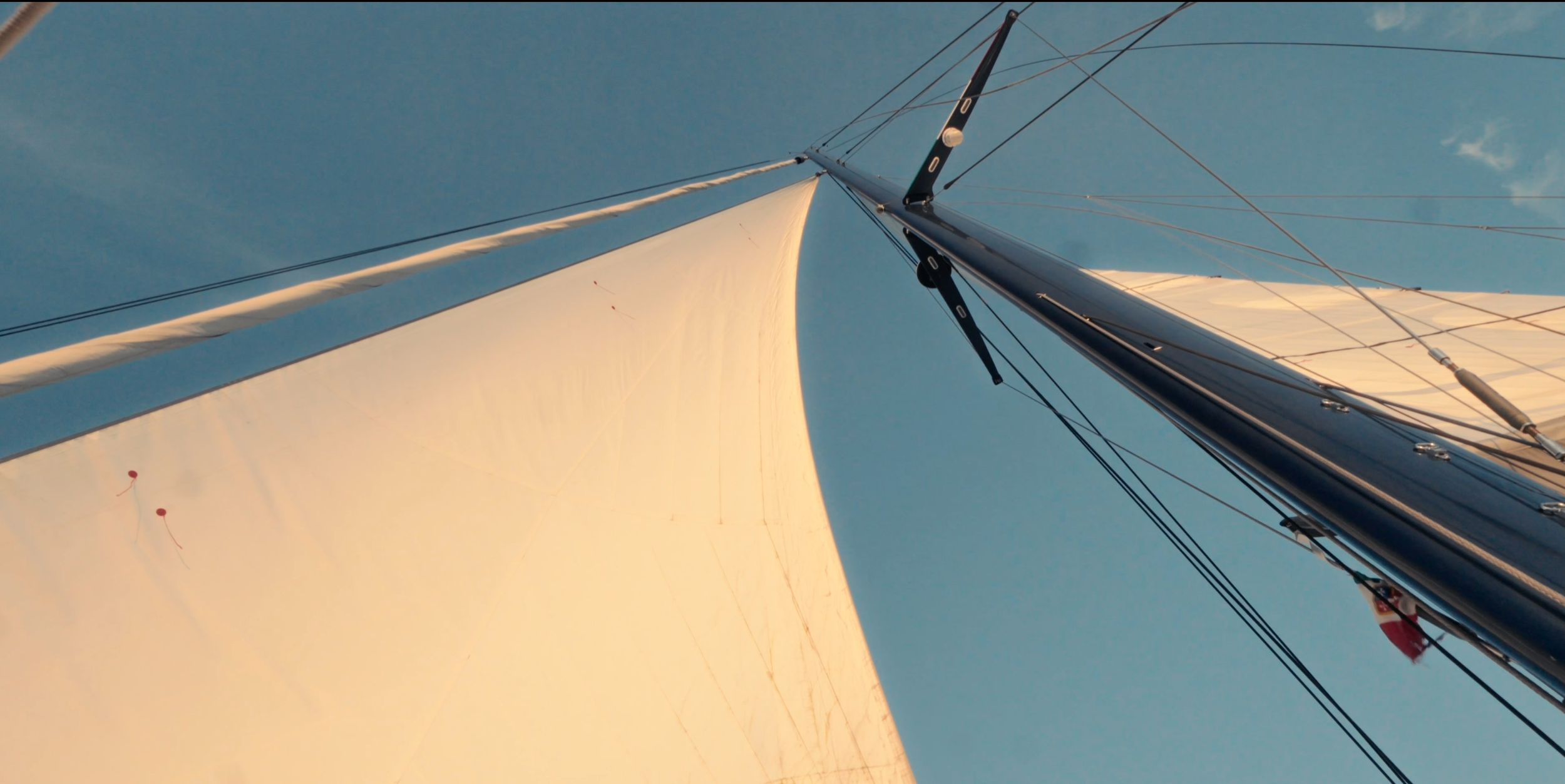 View from the deck of a sailing boat showing the mast, sails, and rigging against a blue sky, with warm sunlight illuminating the sails. LYFE AT SEA