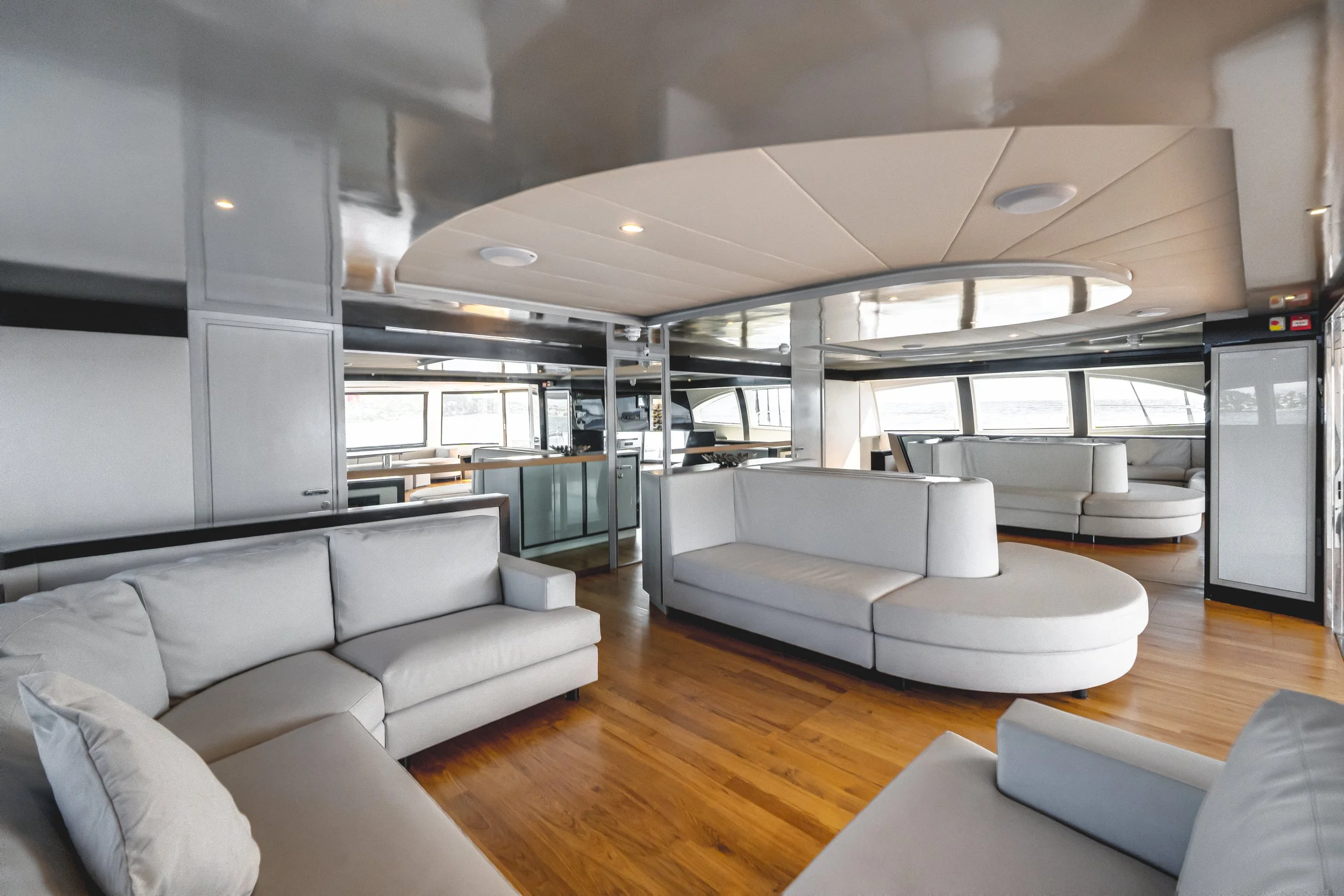 Interior of a modern yacht or boat with white leather sofas, wooden floors, large windows, and a curved ceiling with recessed lighting. LYFE AT SEA
