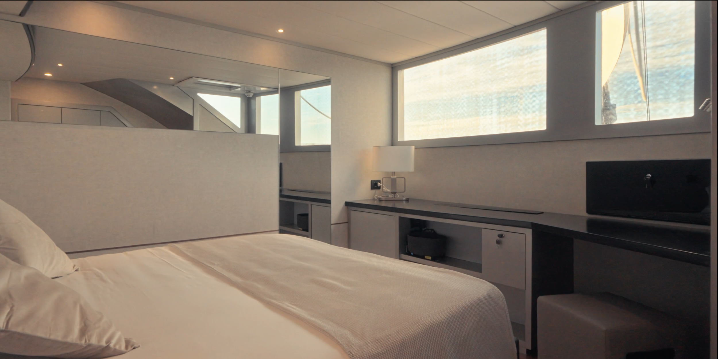 A hotel room with a bed, a desk with a lamp, a small fridge, and large windows letting in natural light. LYFE AT SEA