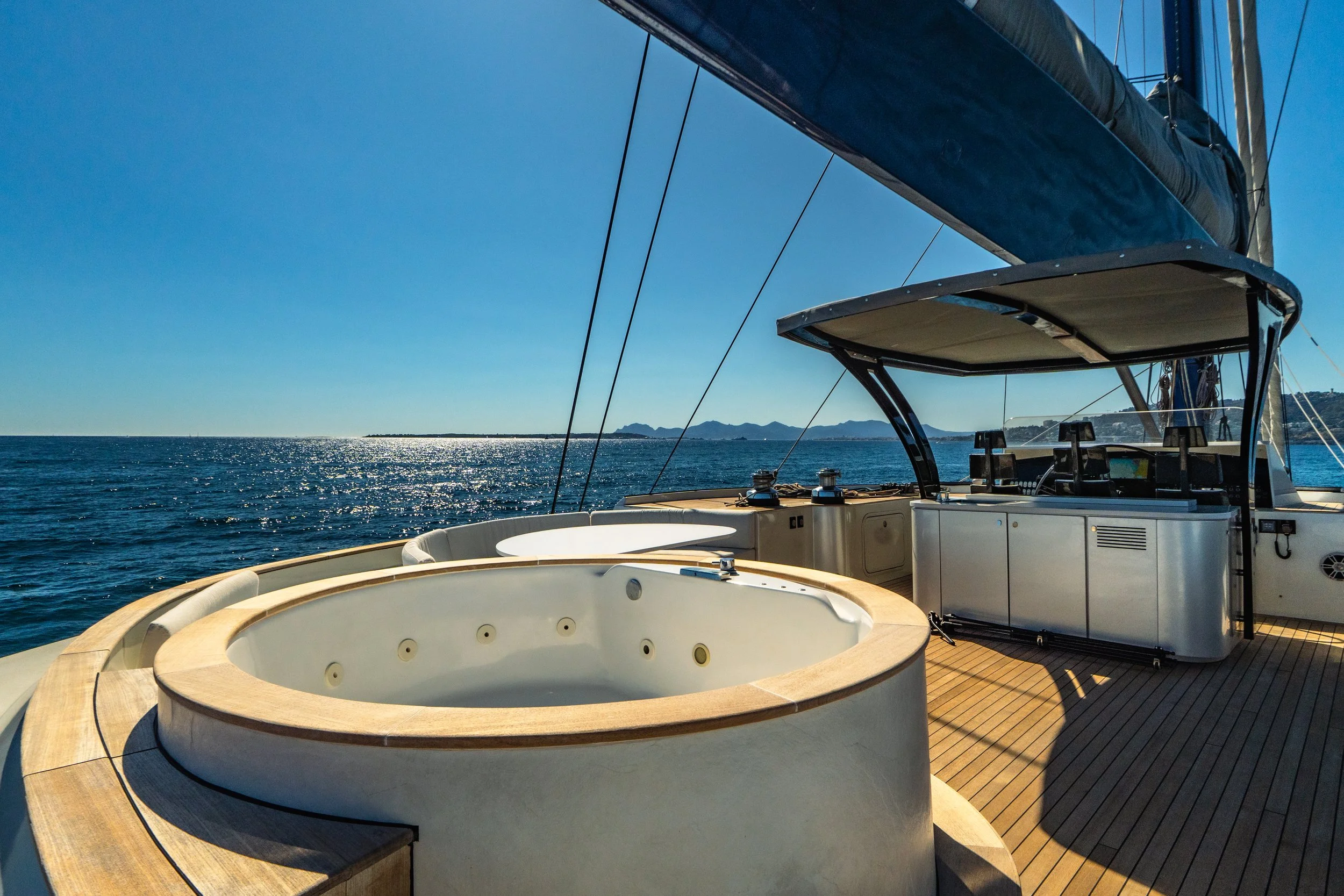 View of a yacht deck with a large circular hot tub, seating area, and a shaded helm station, overlooking the ocean with distant landforms under clear blue skies.