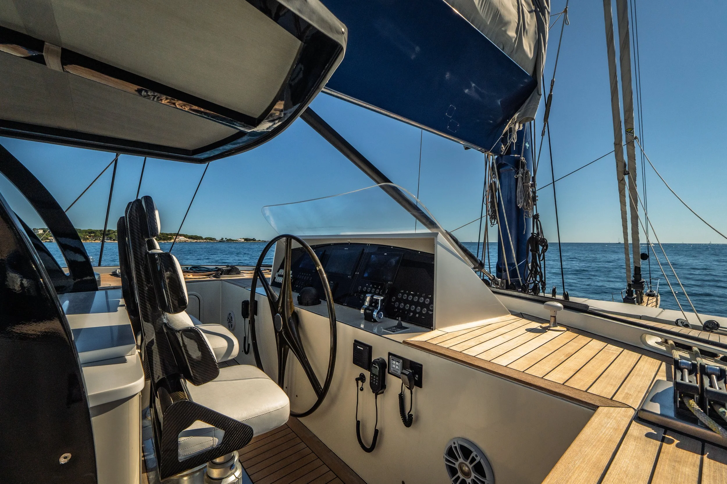 View of a yacht's cockpit with black and white seats, control panels, wiring, and a wooden deck, overlooking calm waters and a distant shoreline under a clear blue sky.