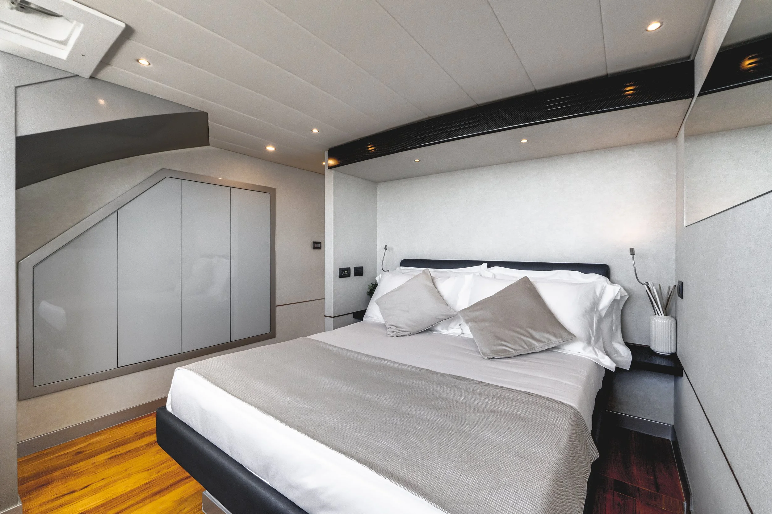 Modern bedroom with a large bed, white bedding, multiple pillows, wood flooring, built-in wardrobe, bedside table with a white vase, and ceiling lights. LYFE AT SEA