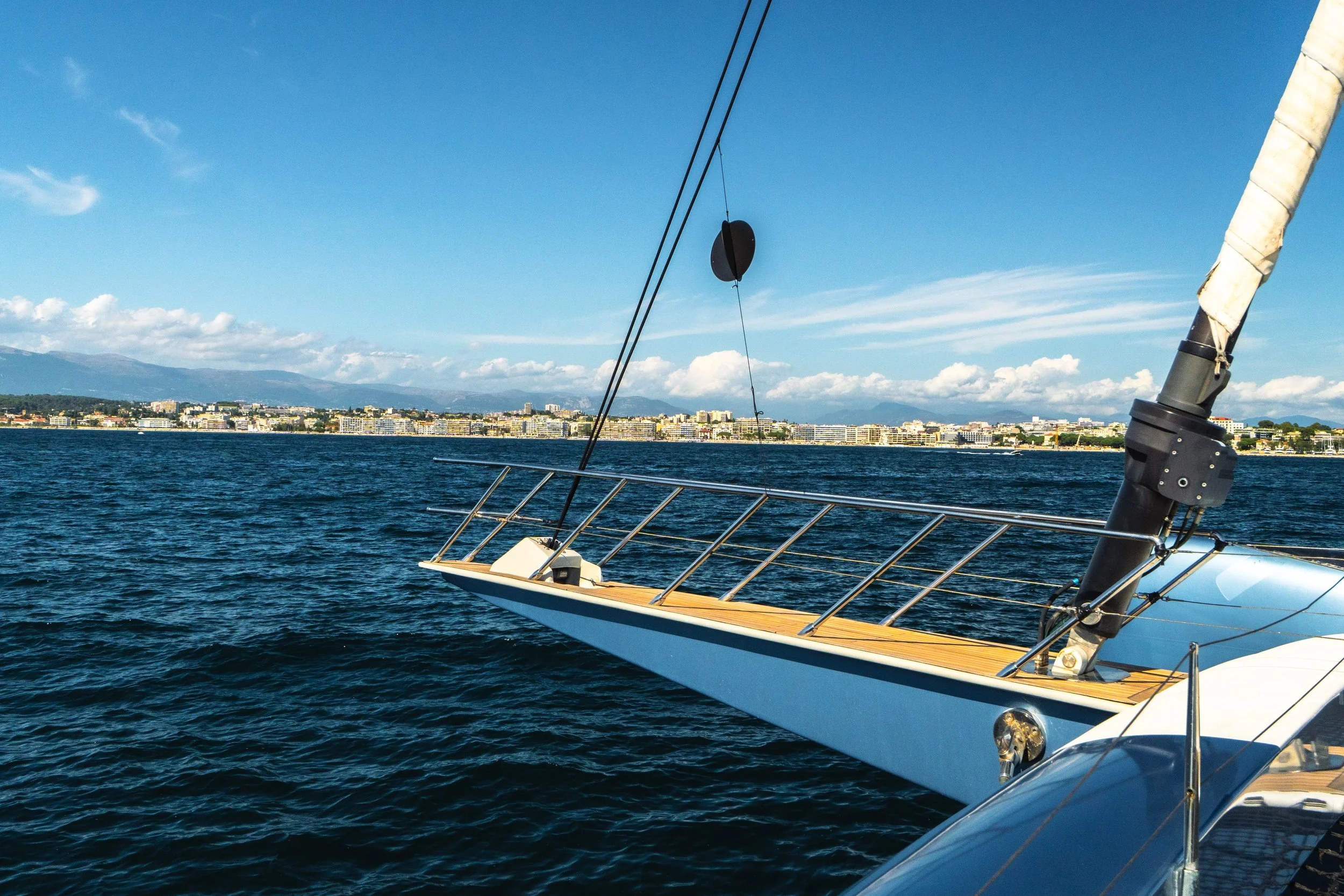 View from a sailboat on the water, showing the bow and rigging, with a city skyline and mountains in the background under a partly cloudy sky. LYFE AT SEA