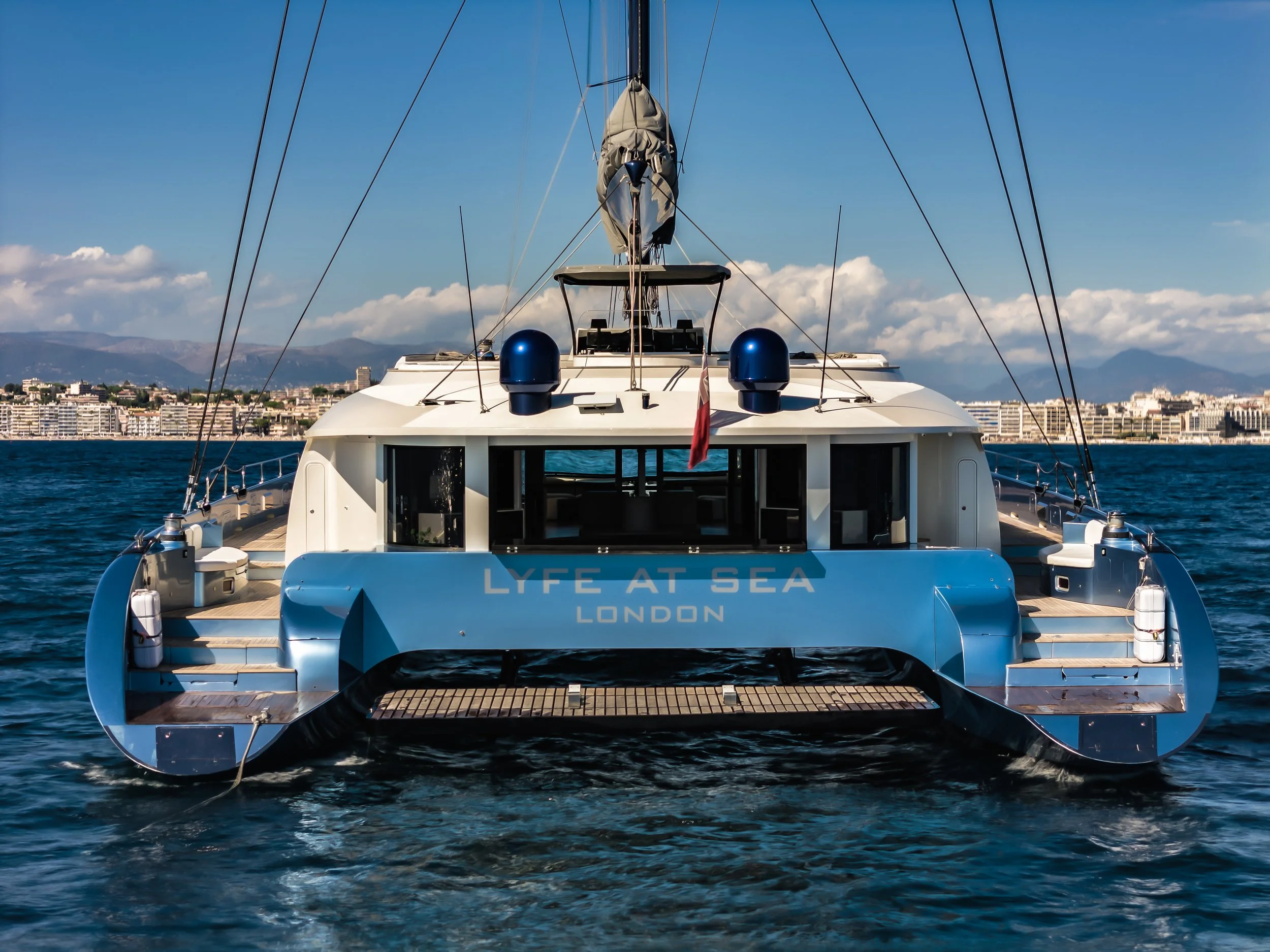 The back view of a large white yacht with the words "LYFE AT SEA LONDON" on the stern, sailing on deep blue water with a cityscape and mountains in the background, under a clear sky. LYFE AT SEA