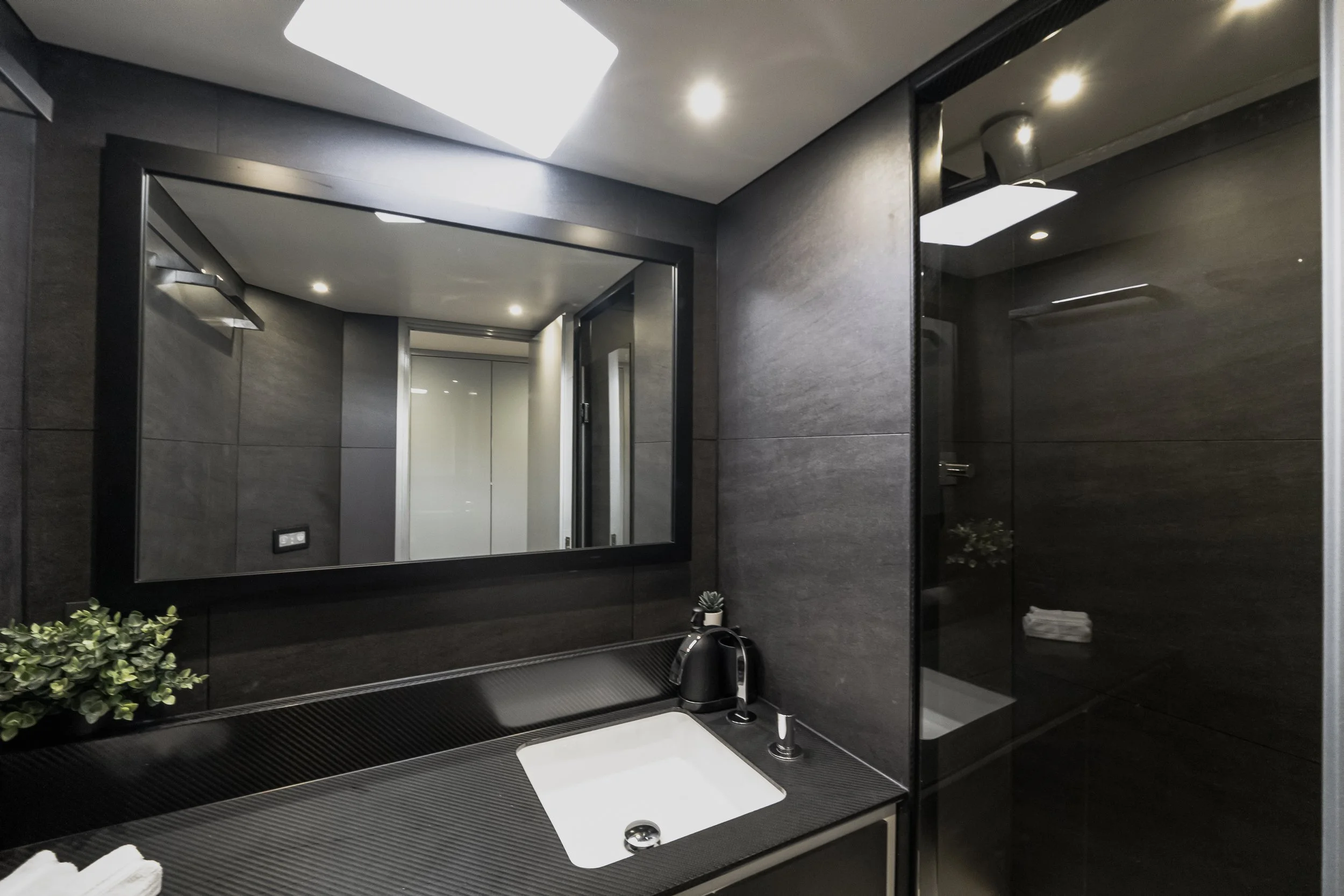 A modern bathroom with dark gray tiles, a black-framed mirror, a white sink, and a black countertop. There is a small plant and folded towels on the counter, with a mirror and reflection of the ceiling lights.