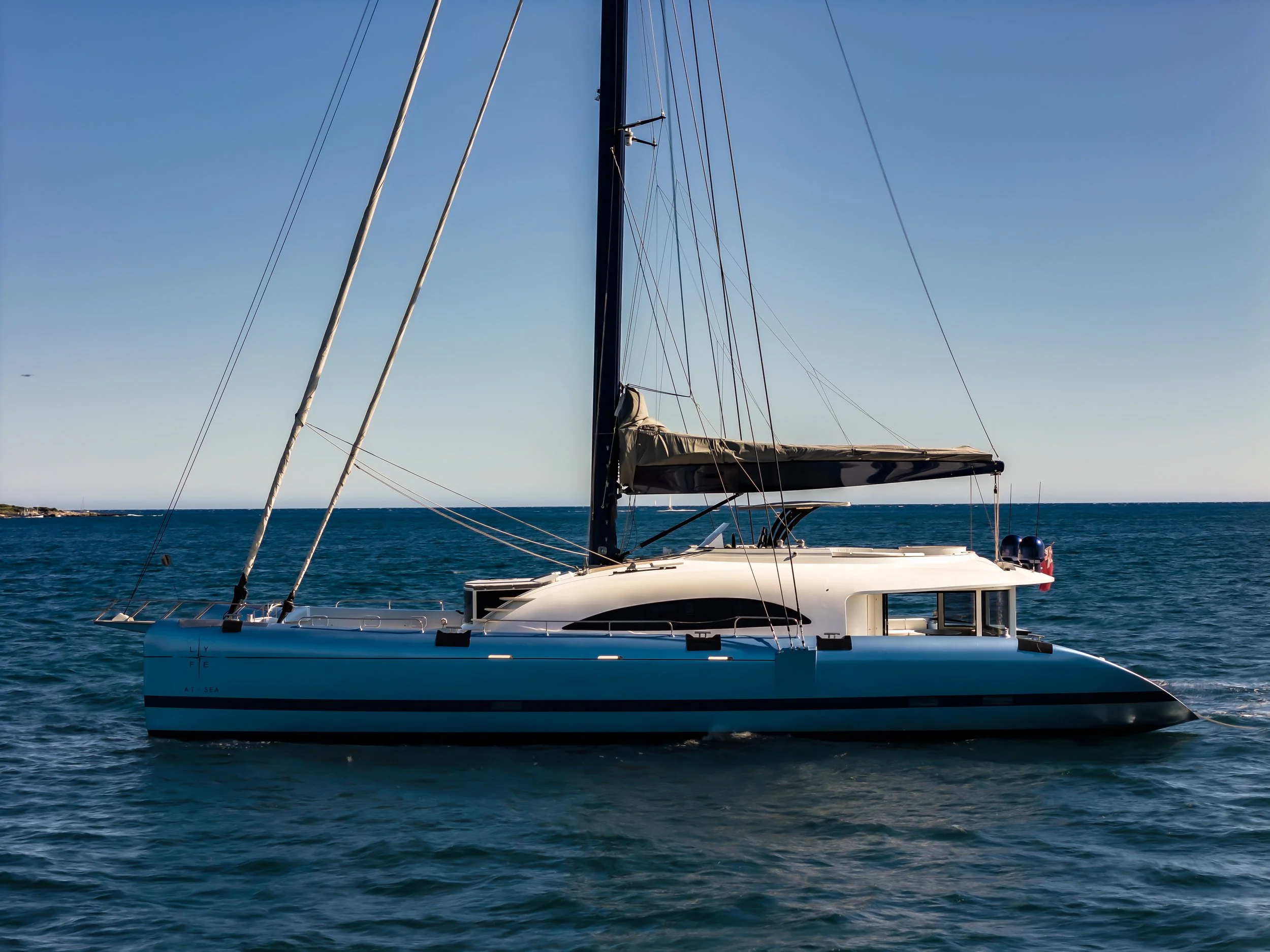 A sailboat on the ocean with a clear sky, multiple ropes and a large sail, and land visible in the background. LYFE AT SEA