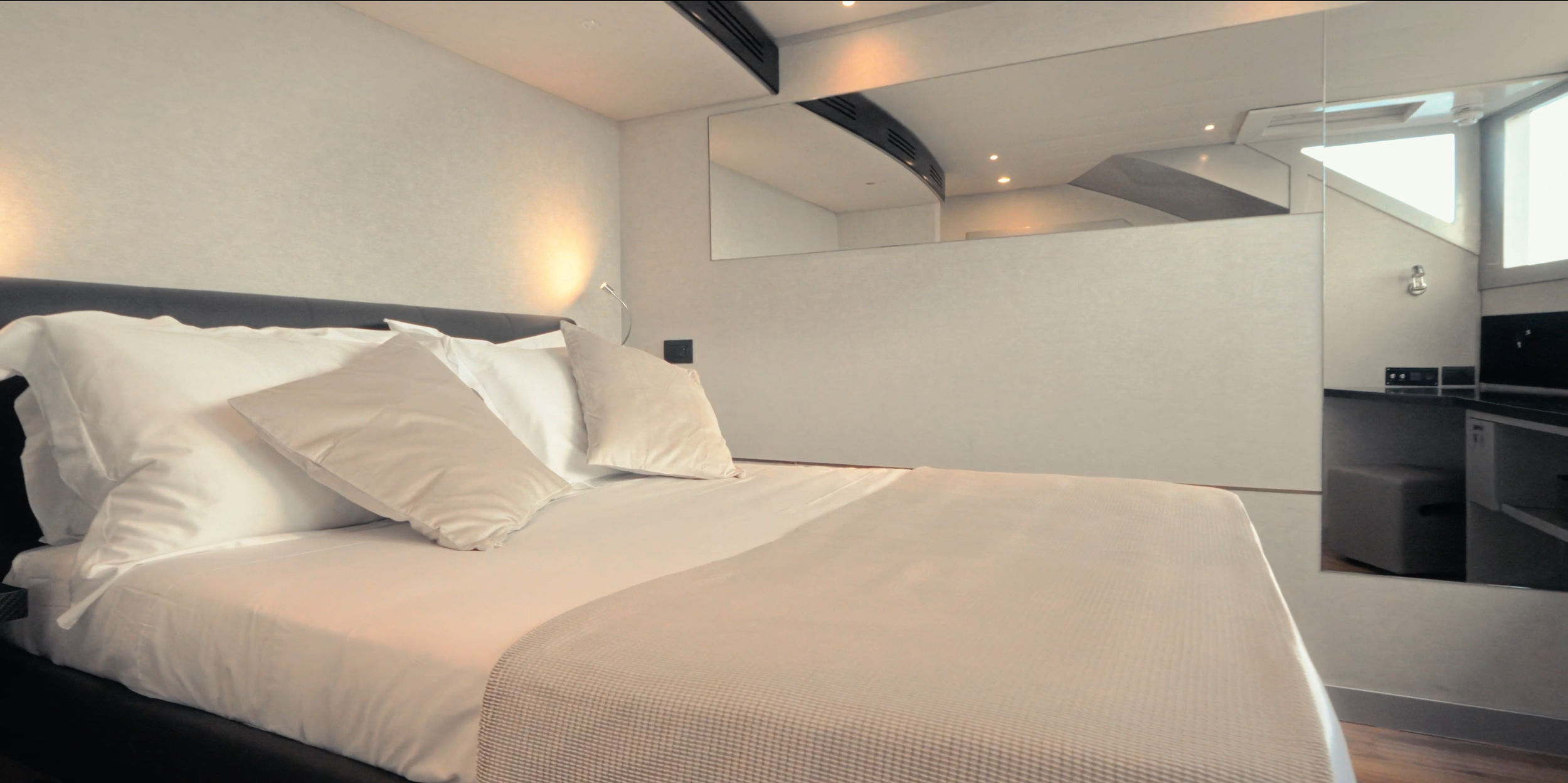Modern hotel room with a neatly made bed, white bedding, and pillows. The room features a minimalist design with neutral colors, a large mirror on the wall, and a window allowing natural light. LYFE AT SEA