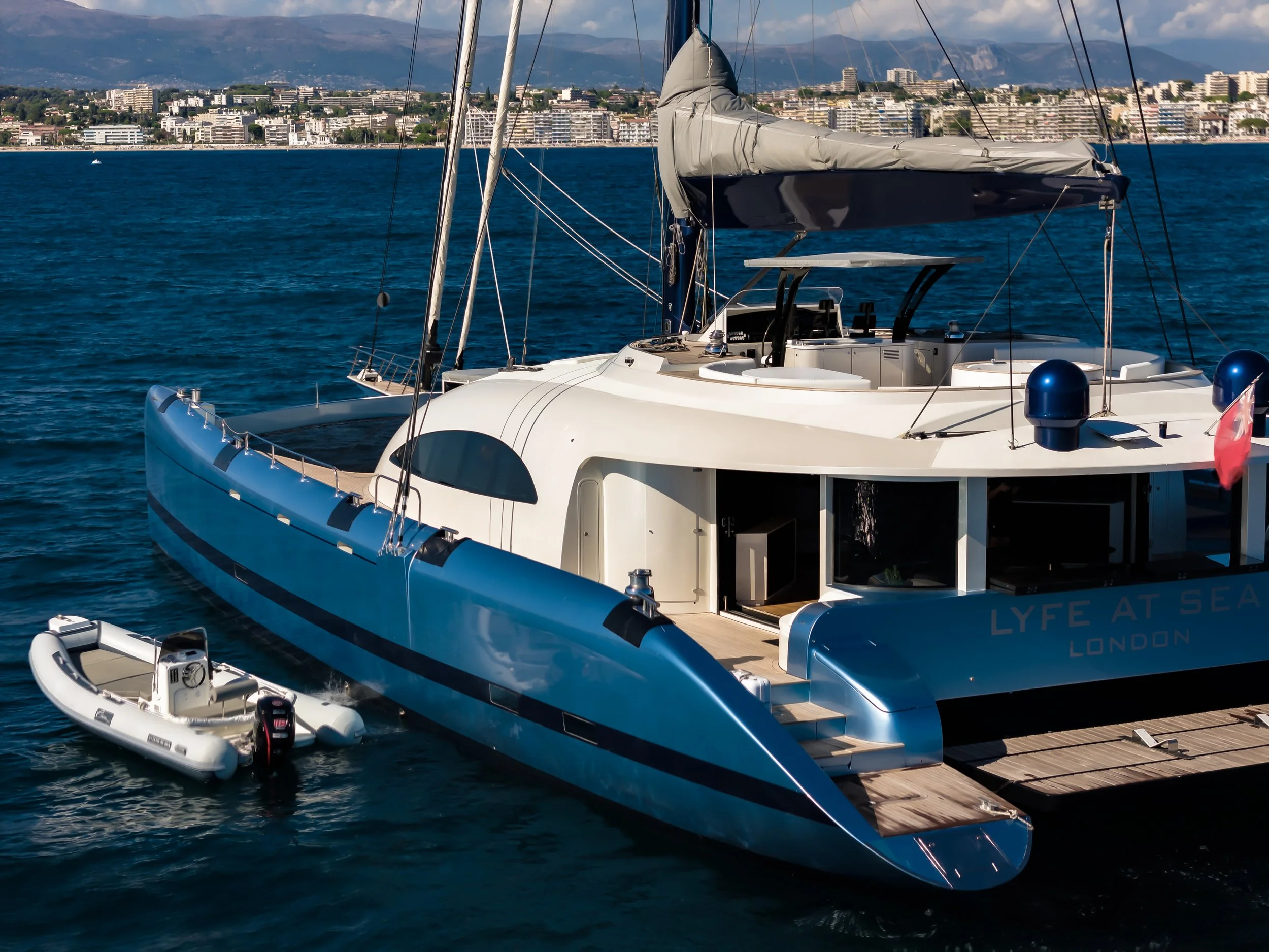 A modern white sailboat docked at a pier with a small inflatable boat attached. In the background, there is a cityscape along the coastline with multiple buildings, and distant mountains under partly cloudy sky. LYFE AT SEA