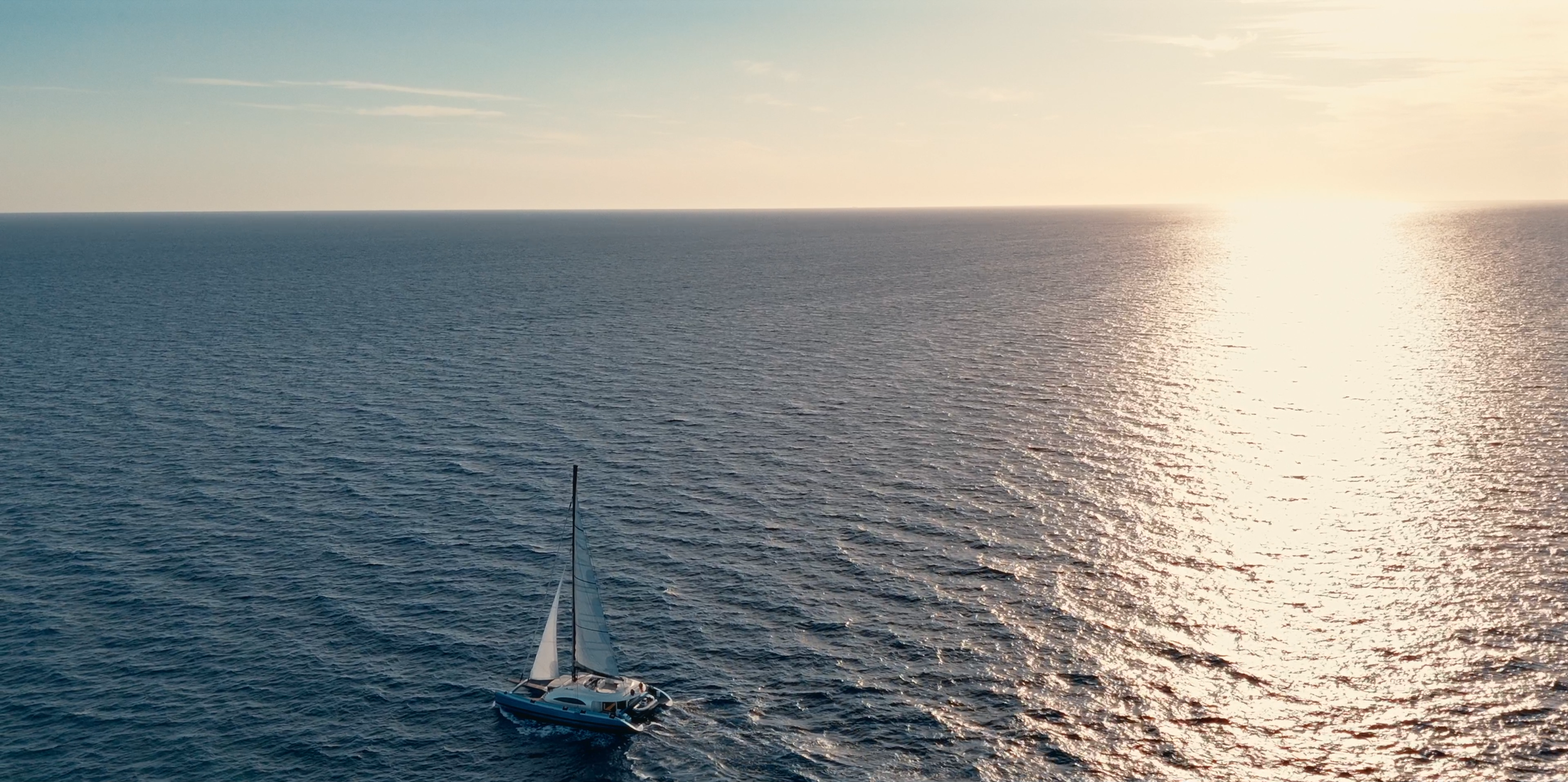 A sailboat with white sails navigating on calm open water during sunset, with the sun reflecting off the ocean surface.