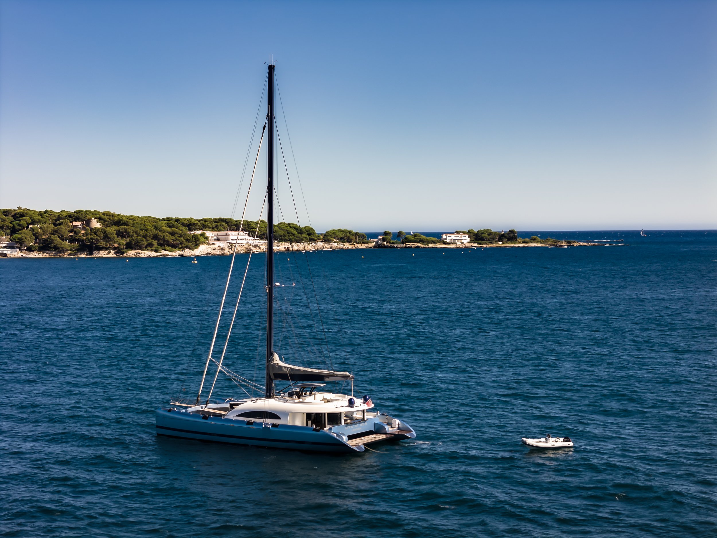A sailboat in a large body of water near a coastline with green trees and houses, under a clear blue sky. LYFE AT SEA