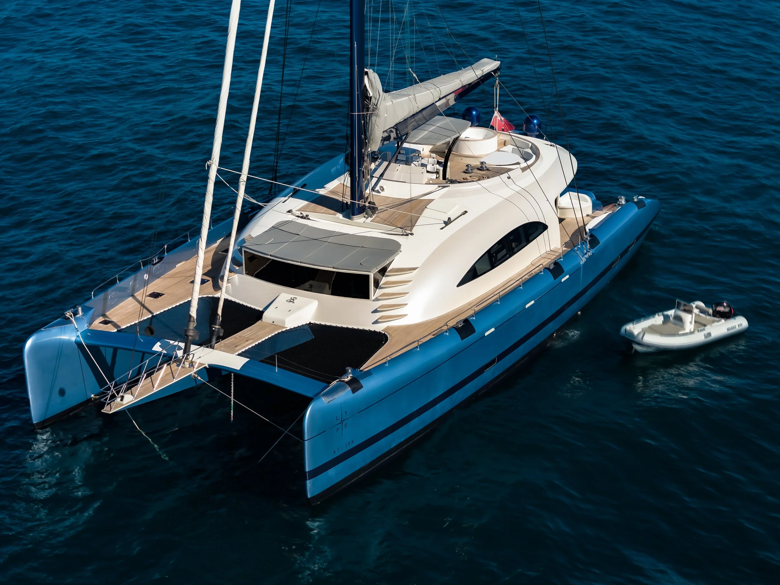 A white luxury sailboat with a wooden deck sailing on the open ocean, featuring a covered cockpit, multiple blue fenders, and a small inflatable boat attached. LYFE AT SEA