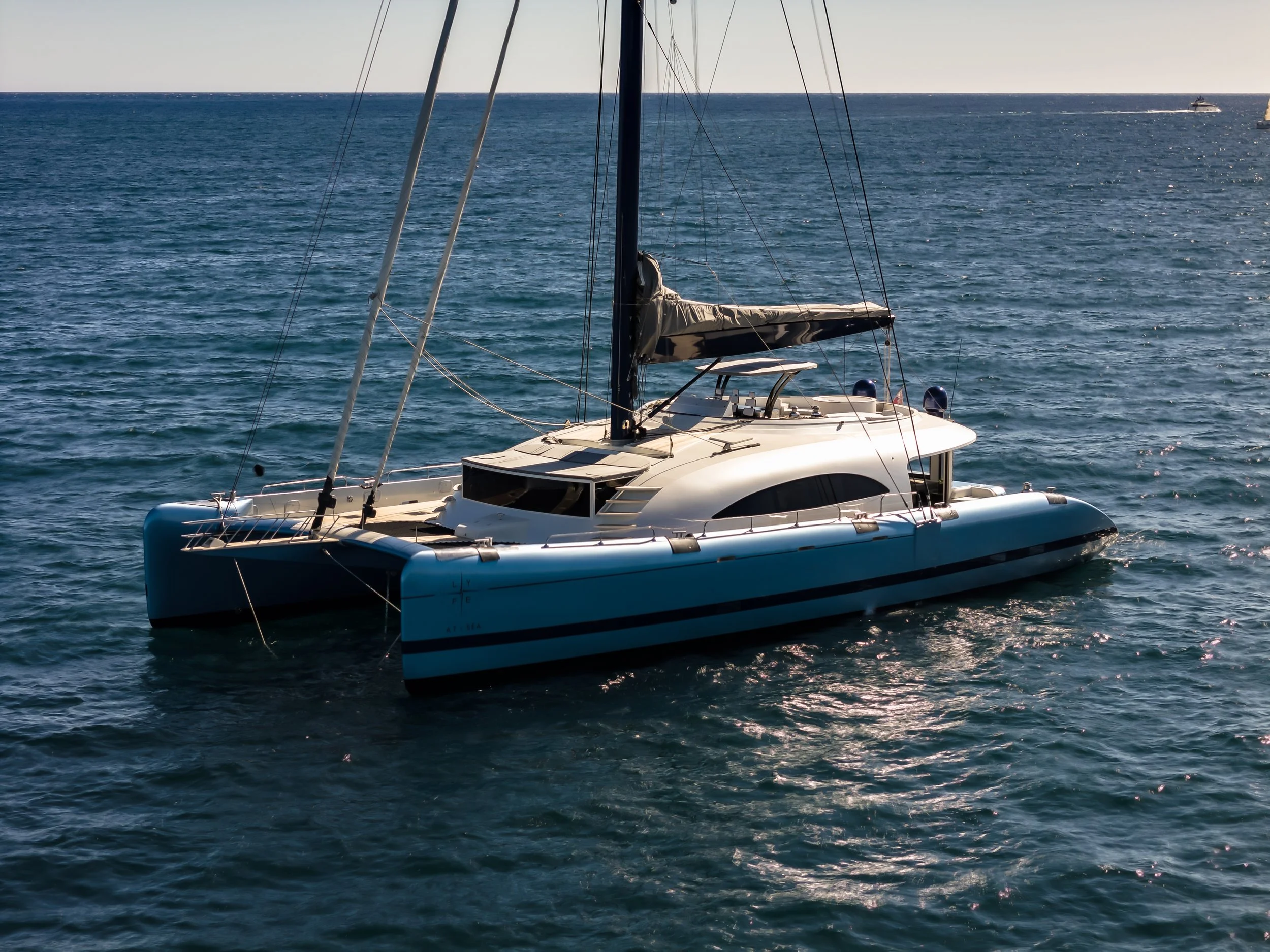A white sailing yacht in open water with another boat visible in the distant background, under a clear sky. LYFE AT SEA