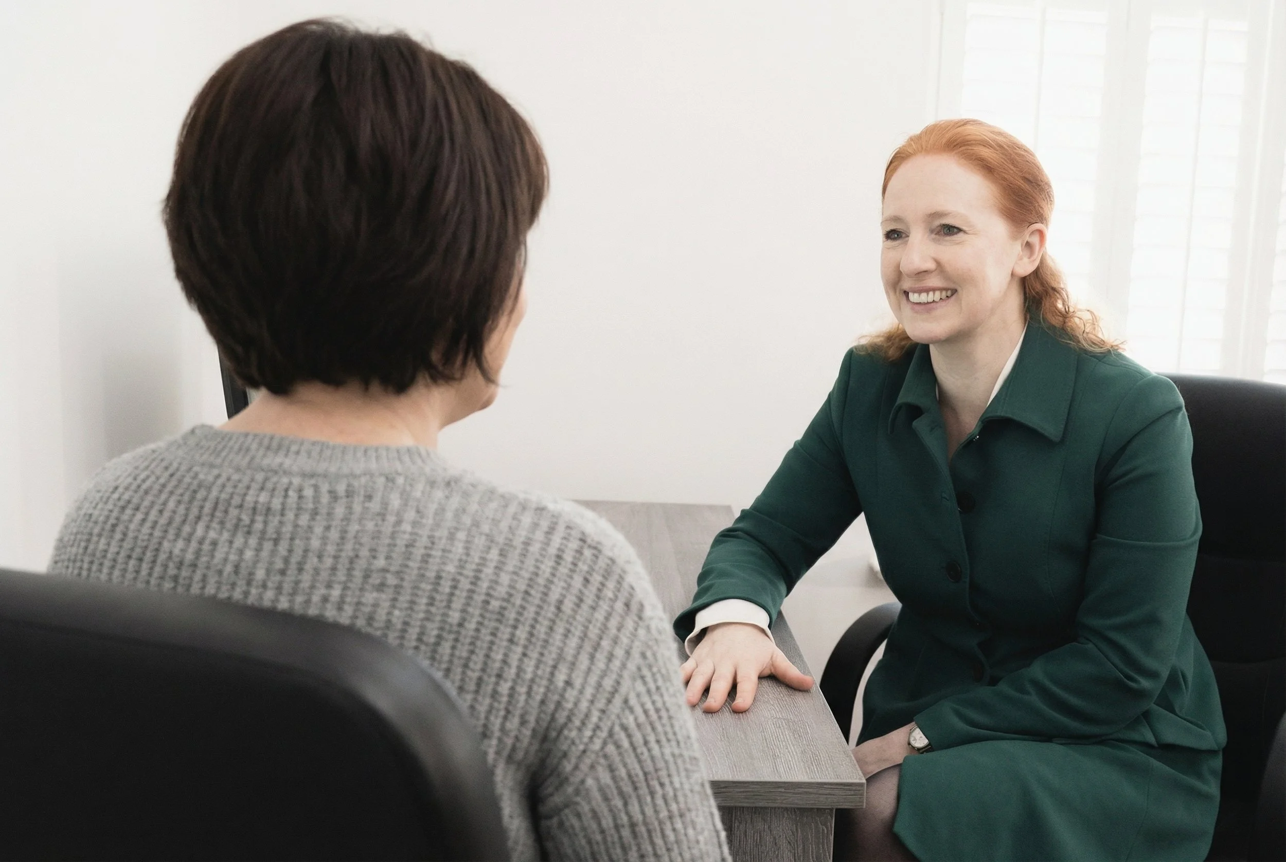 Two women having a conversation in an office, one woman with red hair wearing a green suit, smiling, and the other woman with short dark hair wearing a gray sweater, seen from behind.