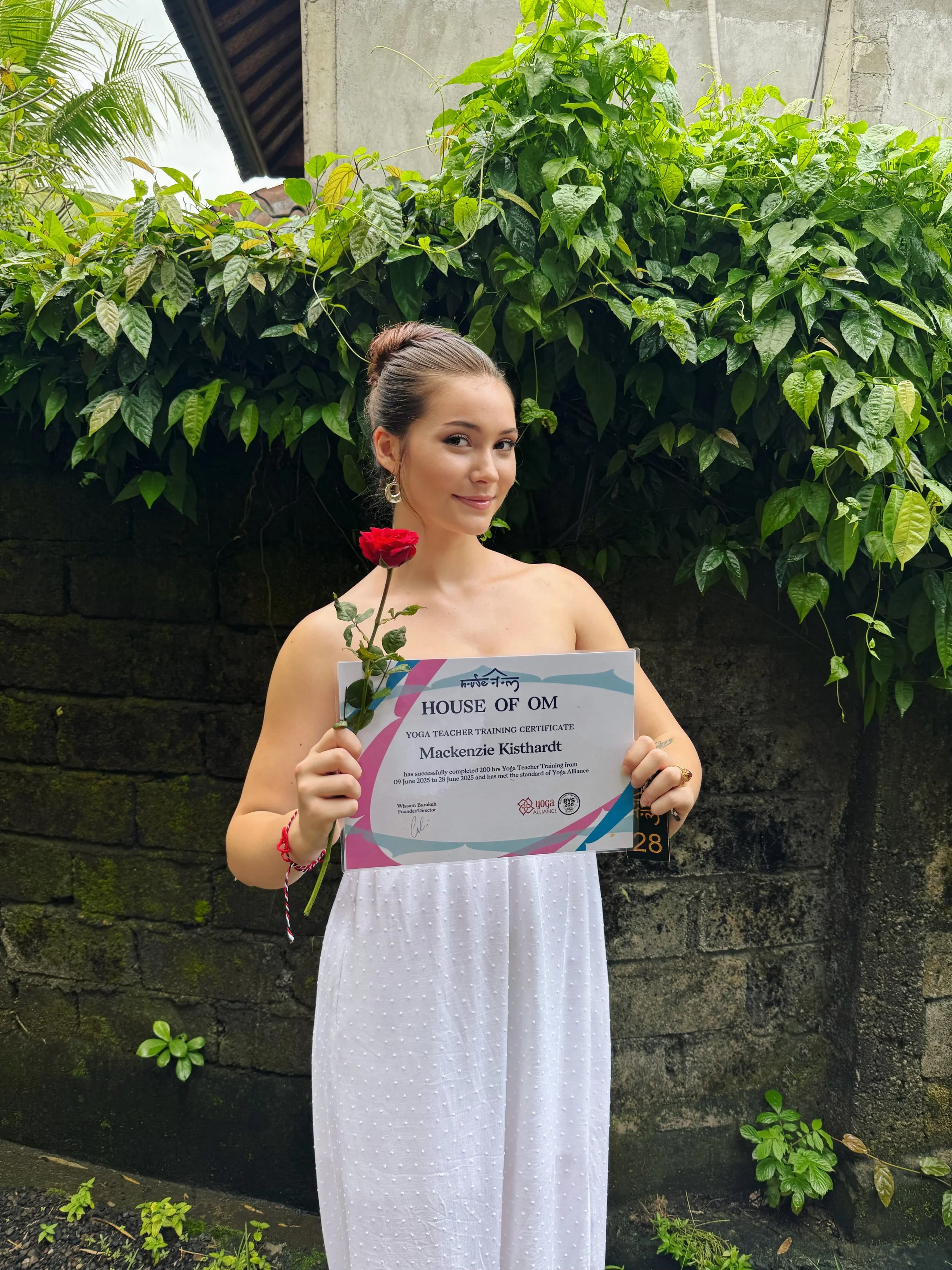 A woman in a white dress holding a certificate and a red rose, standing outdoors against a background of green plants and a stone wall.