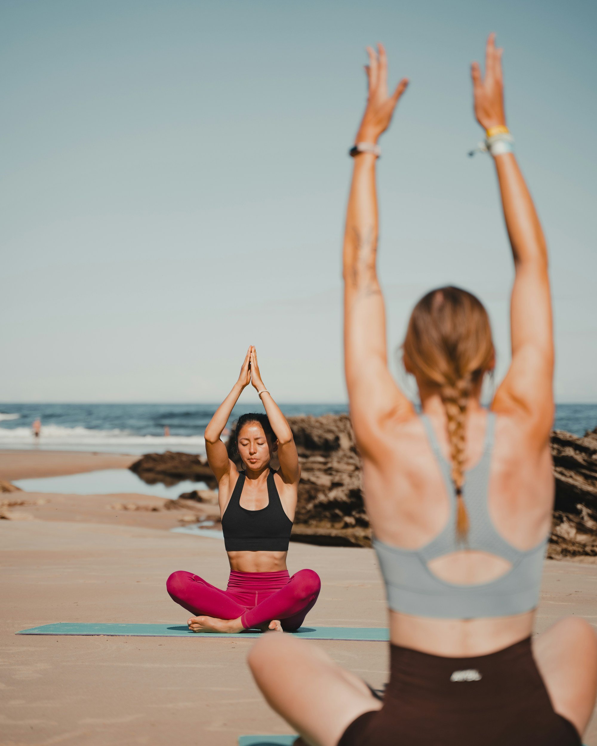 Two women practicing yoga on the beach during sunset, one in the foreground with arms raised and the other in the background sitting cross-legged on a yoga mat with hands pressed together in a prayer position.