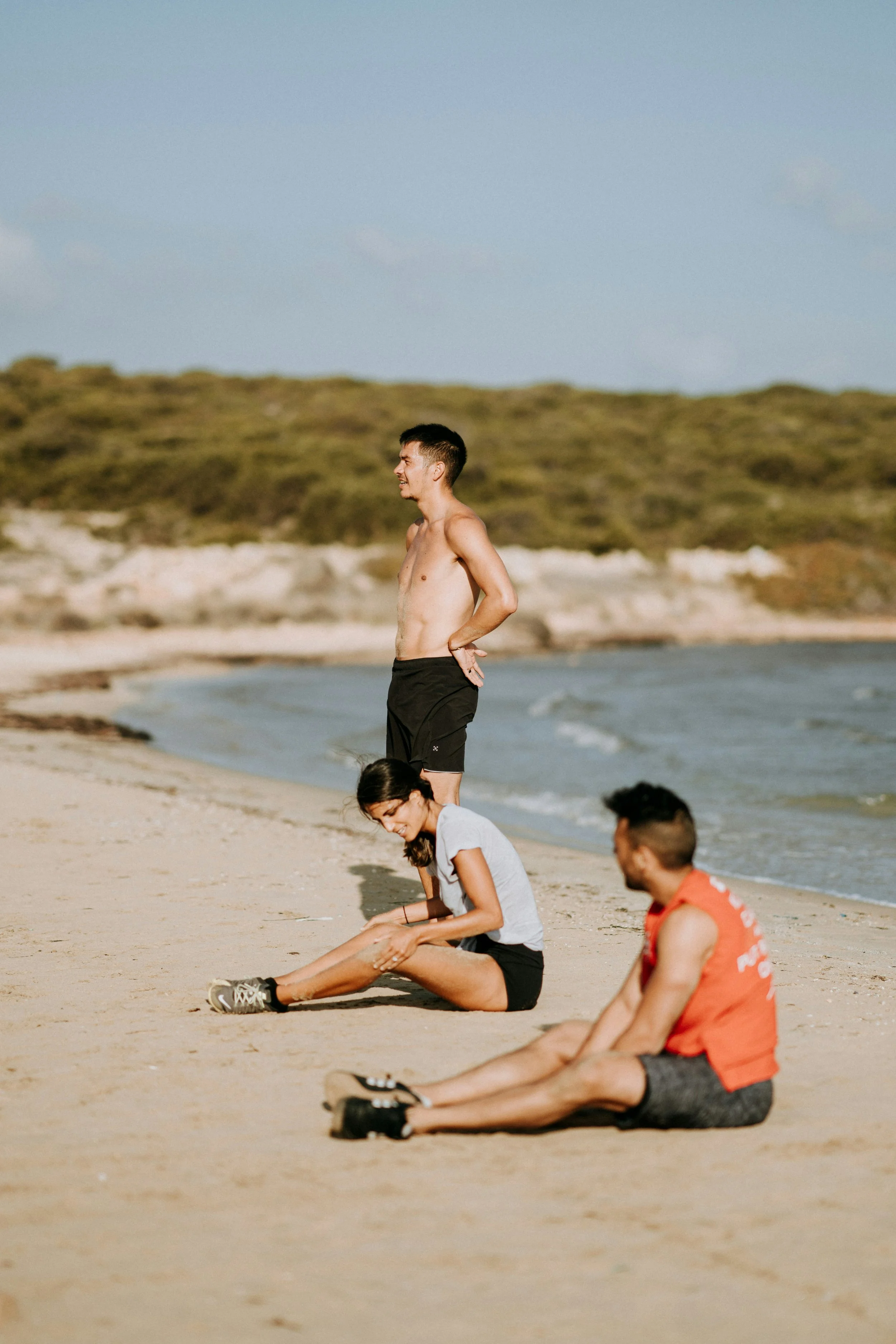 Group of people sitting and standing on the sandy beach near the water's edge, with a man standing shirtless and smiling, and others engaging in relaxing activities.