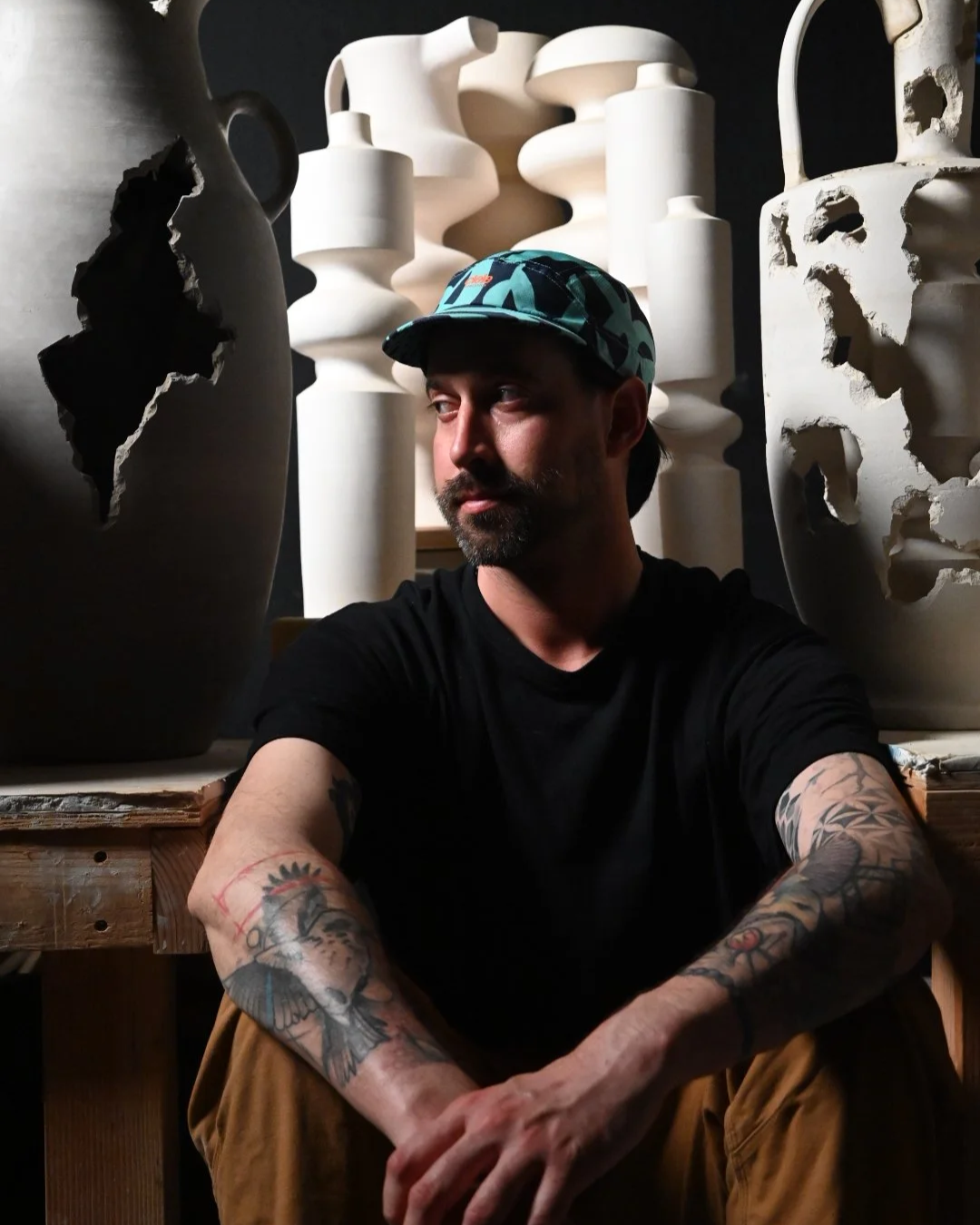 A man with tattoos wearing a black t-shirt and a blue patterned cap sitting in front of large abstract white ceramic sculptures.