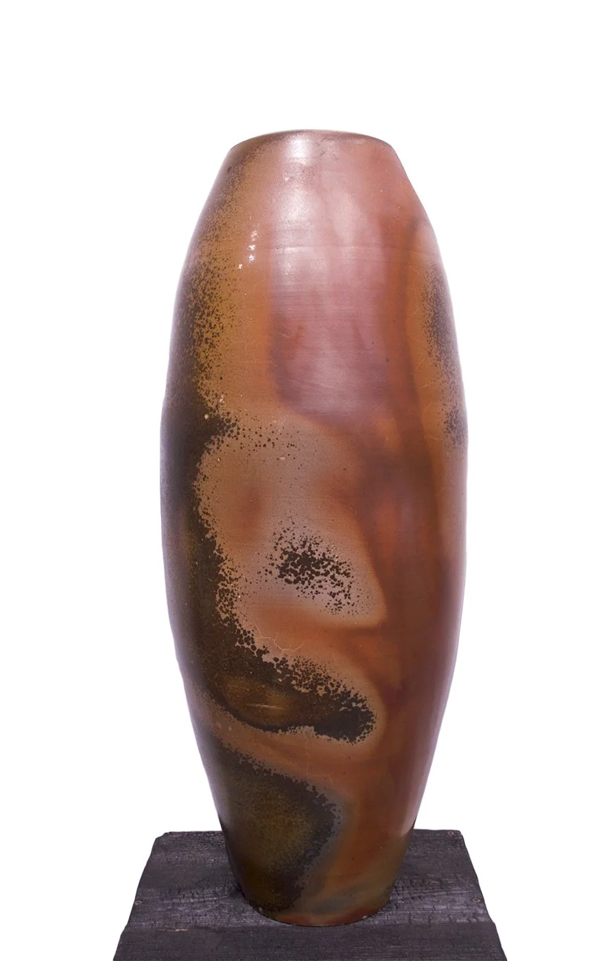 A tall, elongated ceramic vase with a glossy finish, featuring a swirling pattern of earthy tones including browns, reds, and hints of black, mounted on a black wooden base.