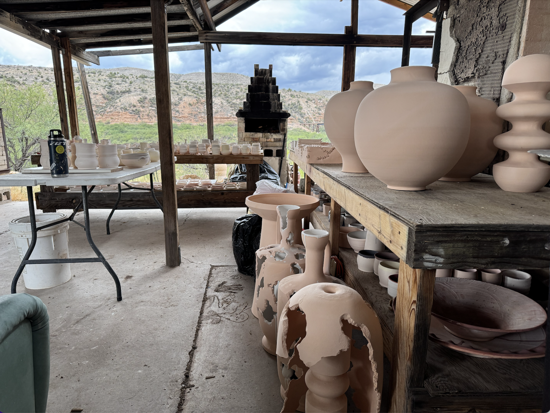 Ceramic art studio with unfinished clay vases and sculptures on wooden shelves, outdoor canyon landscape visible through open walls.