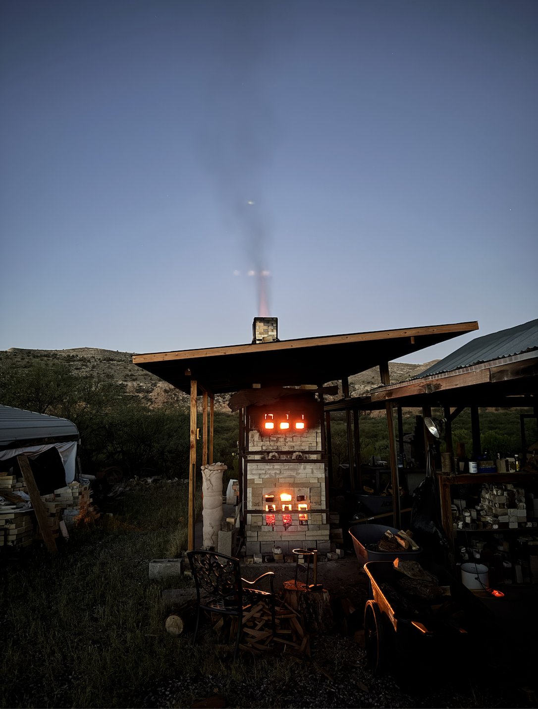 A backyard outdoor fireplace heating bricks at dusk, smoke rising into the clear evening sky, surrounded by miscellaneous construction materials and outdoor furniture.