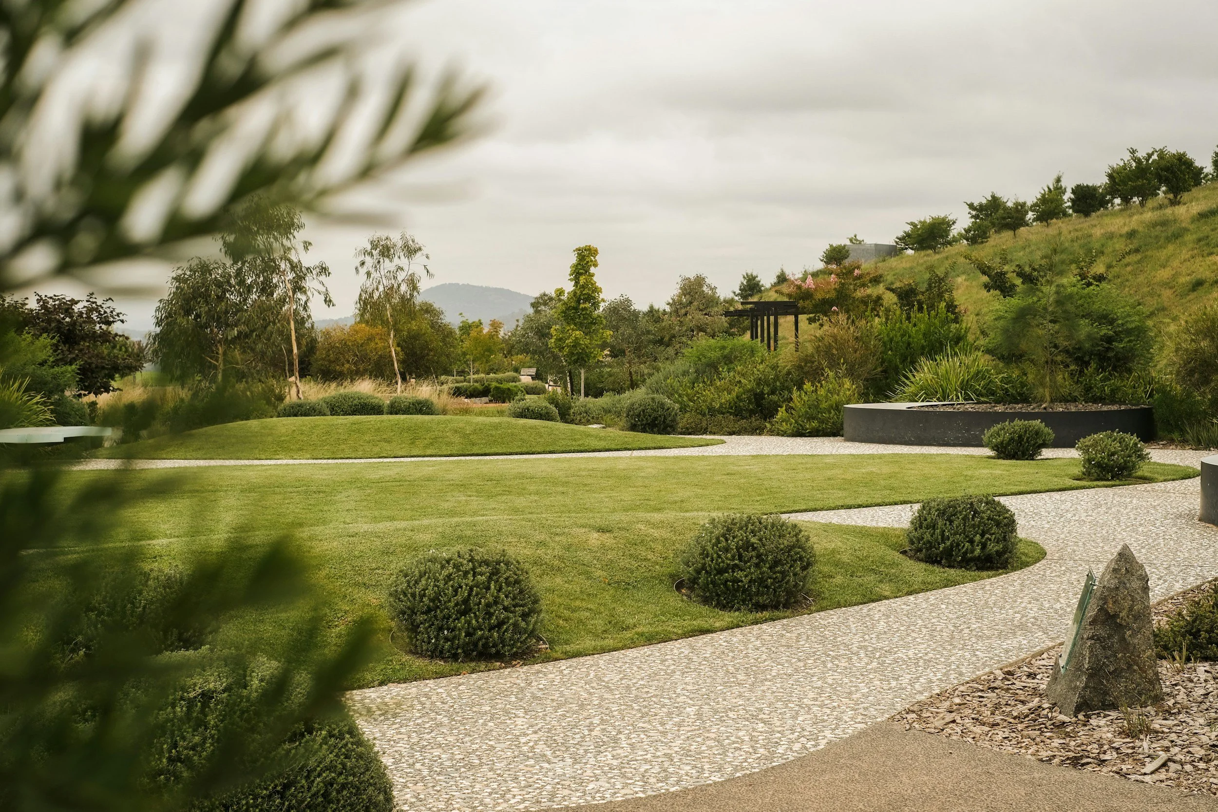 A landscaped garden with green grass, small shrubs, and a paved pathway, with trees on a hillside in the background under a cloudy sky.