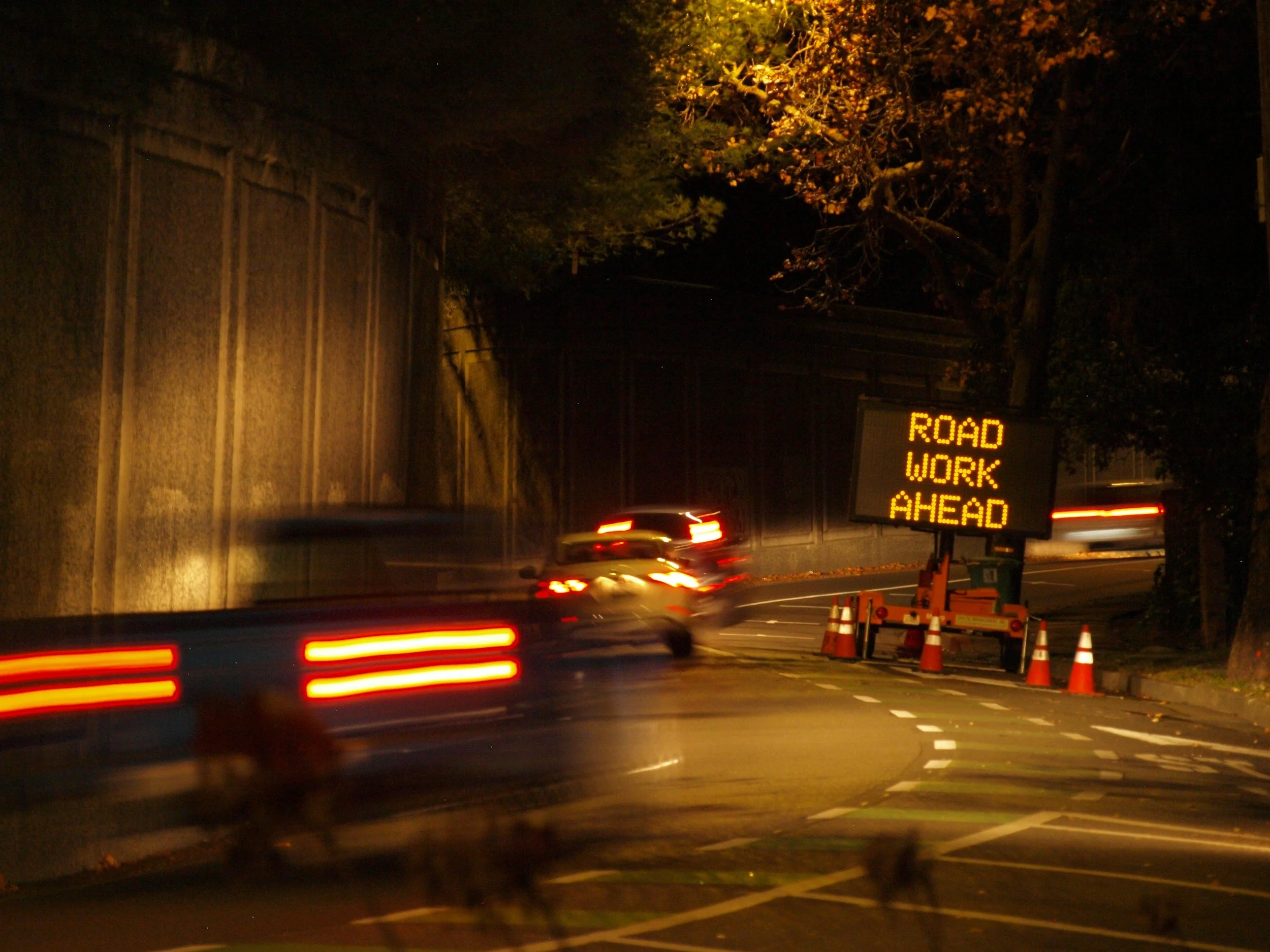 A nighttime scene of a road with cars moving, with motion blur. There is a bright digital sign on the right side displaying 'ROAD WORK AHEAD'. Traffic cones are positioned near the sign. Trees line the street, and a wooden fence is visible on the left side. The scene suggests a road construction zone.