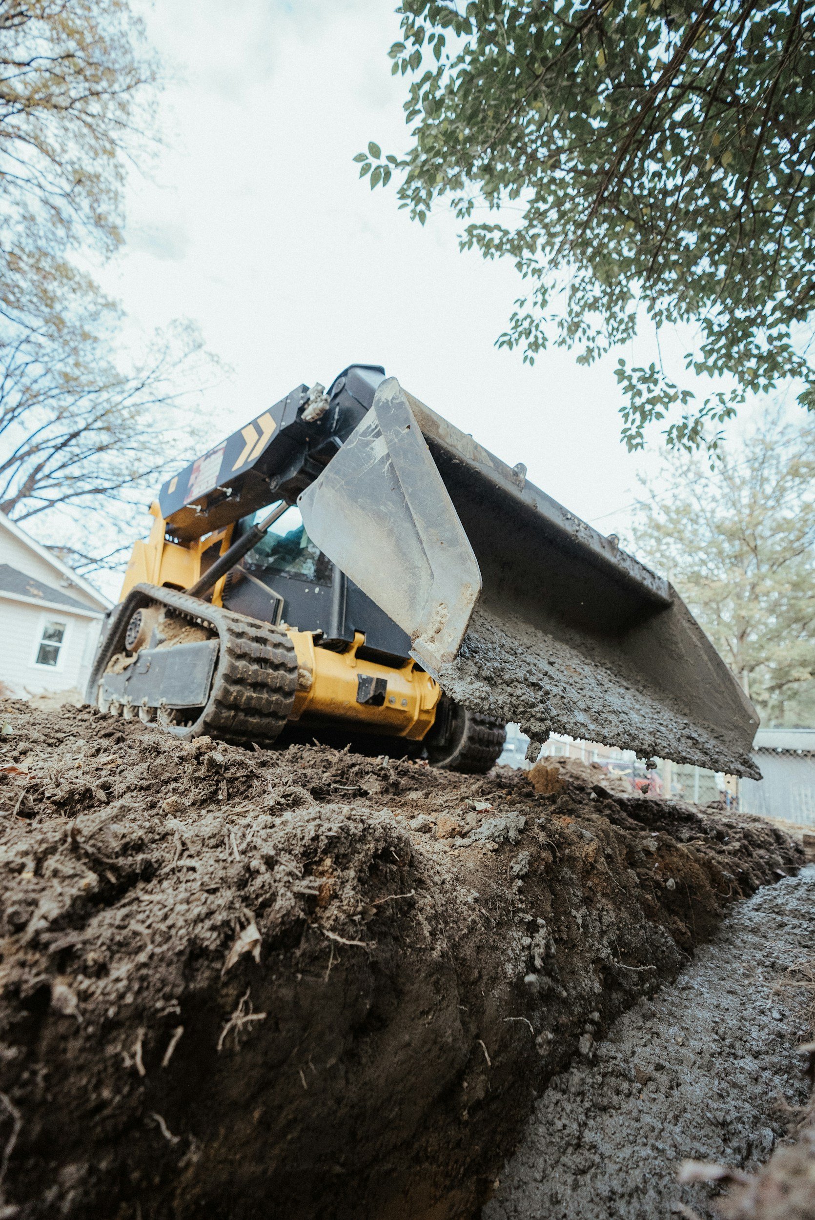 A construction excavator working on a dirt site, with trees and houses in the background.
