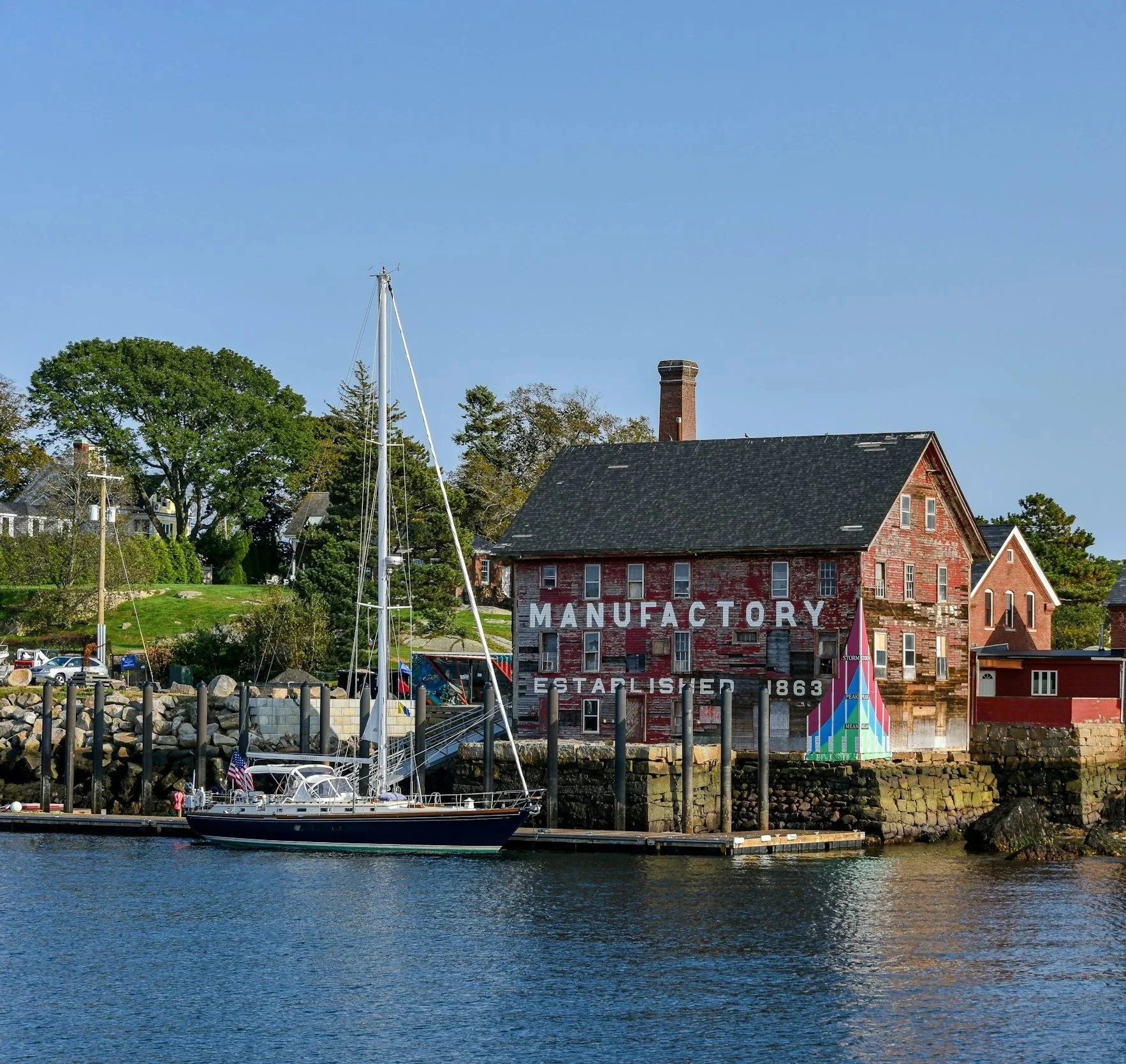 A marina with a sailboat docked in the water, a historic red brick building with a faded sign reading 'MANUFACTURER ESTABLISHED 1863,' surrounded by green trees and houses, under a clear blue sky.