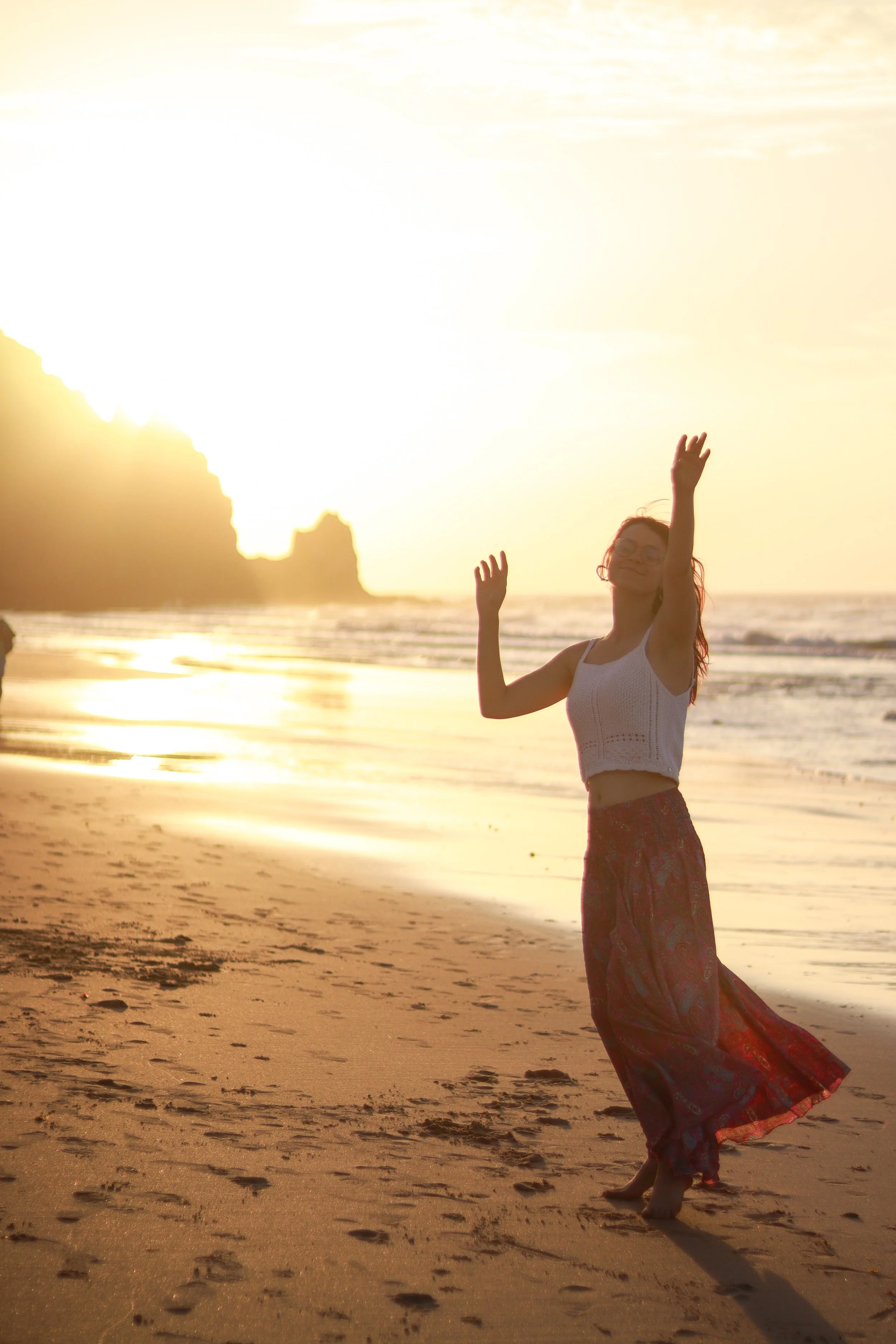 Vrouw die op het strand staat tijdens zonsondergang, met haar armen omhoog en een lange rok, terwijl ze lacht.