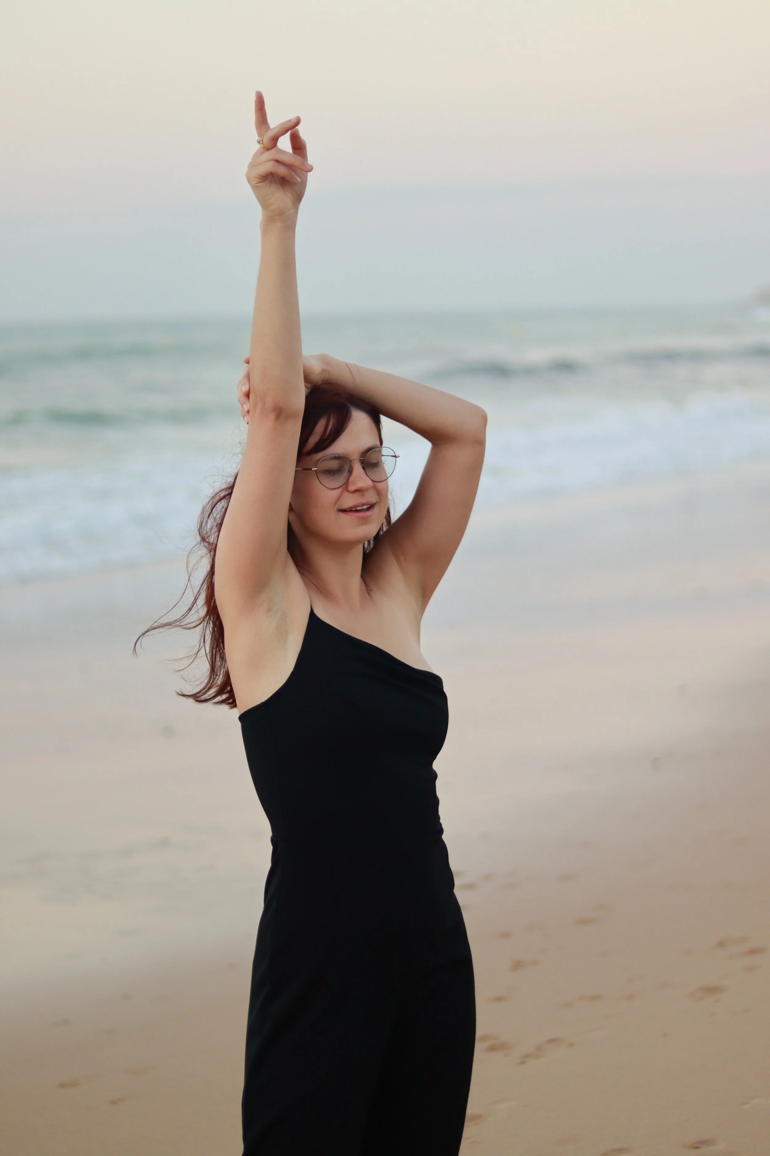 Vrouw in zwart jurkje staat op het strand met de oceaan op de achtergrond, haar ogen gesloten en haar hand in de lucht, alsof ze danst of poseert.