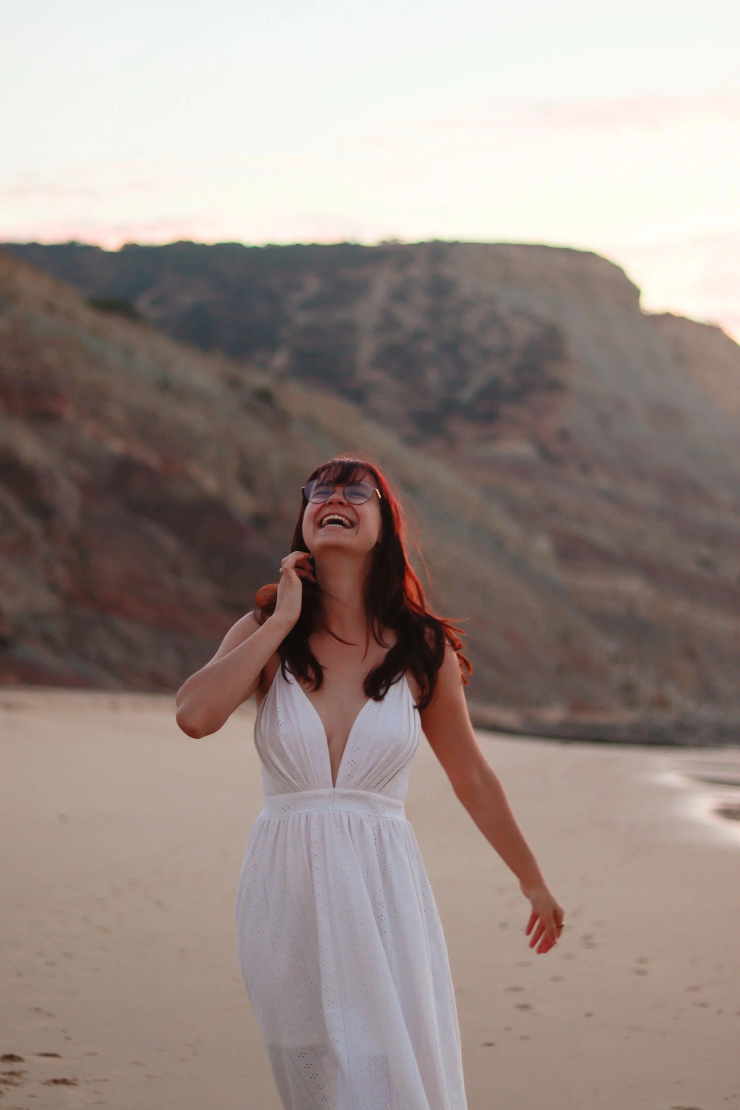 Vrouw in witte jurk lachend op het strand met bergen op de achtergrond tijdens zonsondergang.