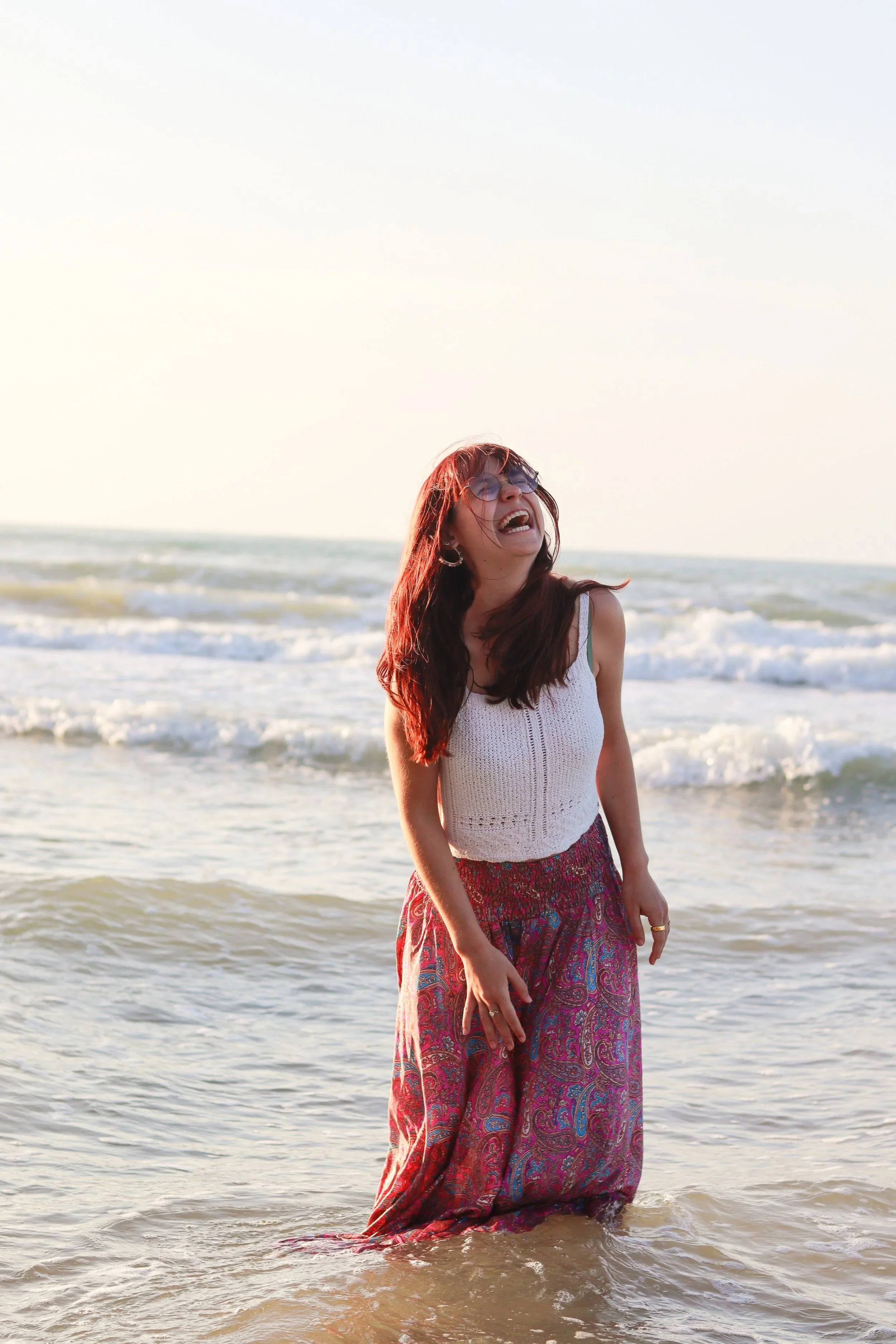 Een vrouw met rood haar, zonnebril en een witte top die in de zee staat, lachend en genietend van de zonsondergang op het strand.