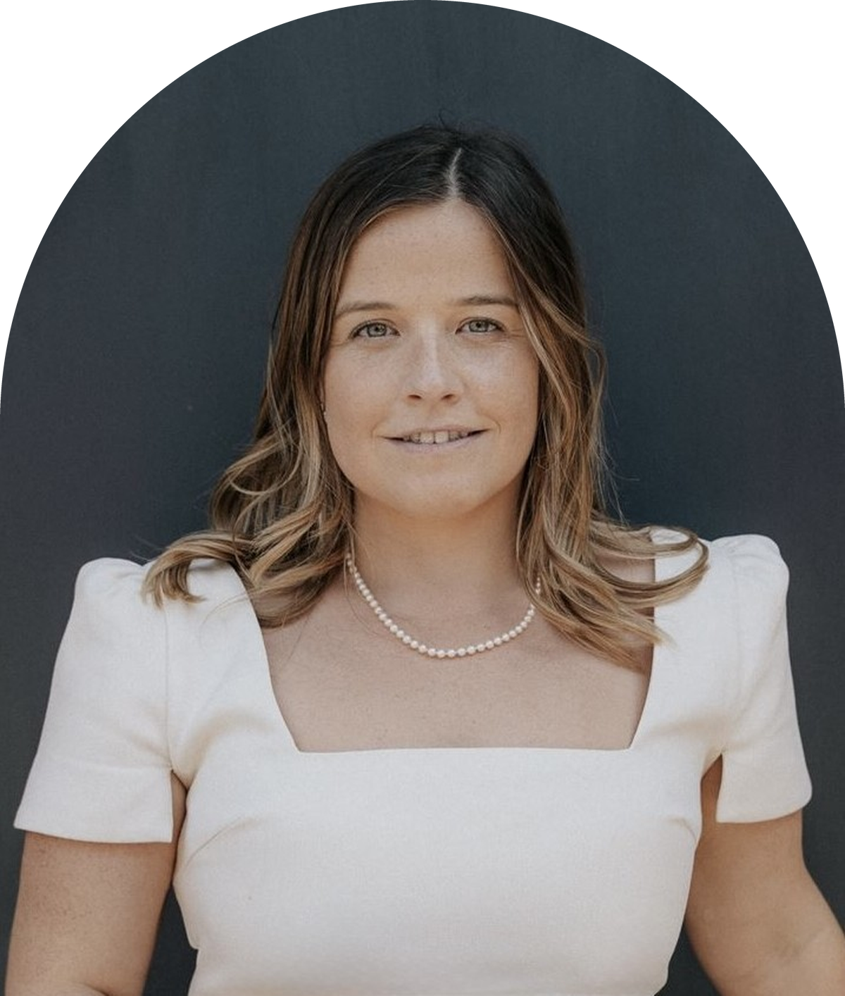 Portrait of a woman with shoulder-length wavy hair, wearing a white top and a pearl necklace, smiling softly against a dark background.