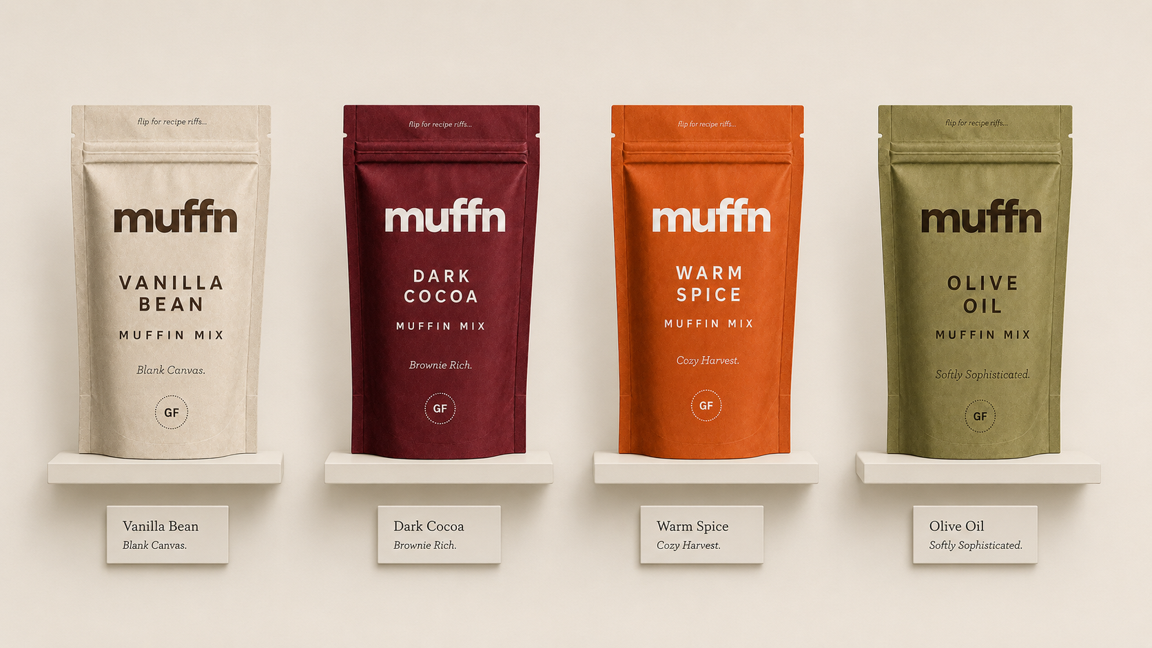 Four colorful bags of muffin mix on a wall shelf, labeled Vanilla Bean, Dark Cocoa, Warm Spice, and Olive Oil, each with a small descriptive tag below.