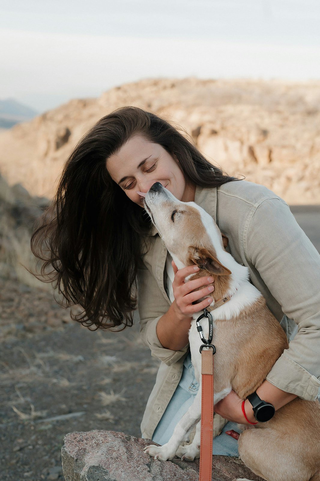 Evelyn Iacono hugging a small brown and white dog outdoors with rocky terrain and mountains in the background.