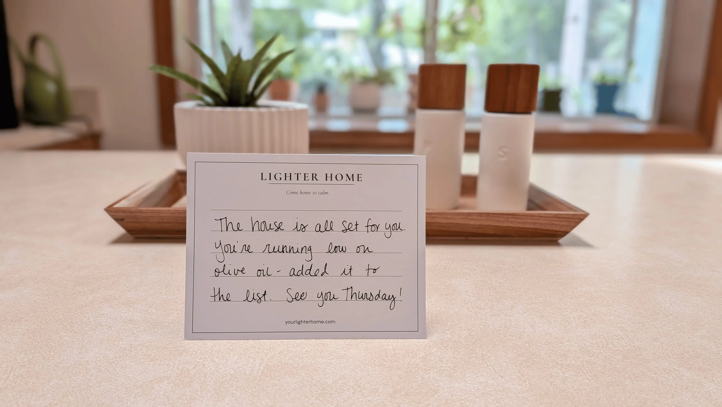 Handwritten note from Lighter Home that reads "House is all set for you. You're running low on olive oil - added it to the list. See you Thursday!" In the background, potted plants on a windowsill, with blurred green trees visible outside the window.