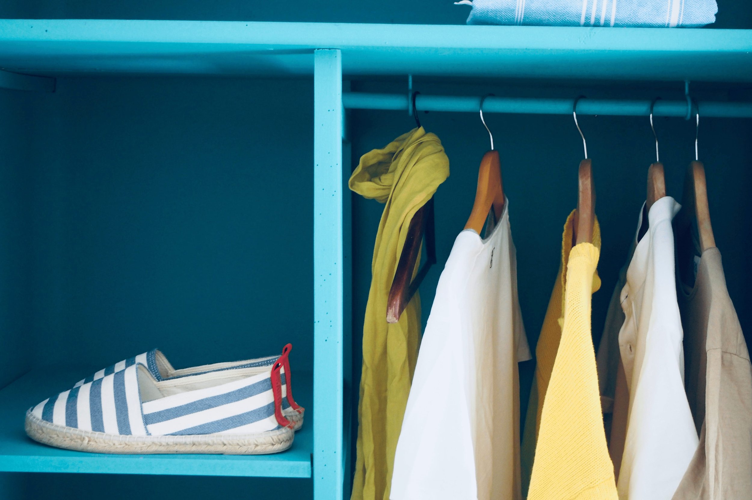 Inside a closet, a blue shelf with a pair of striped canvas shoes, and yellow, white, beige, and brown clothing hanging on wooden hangers.