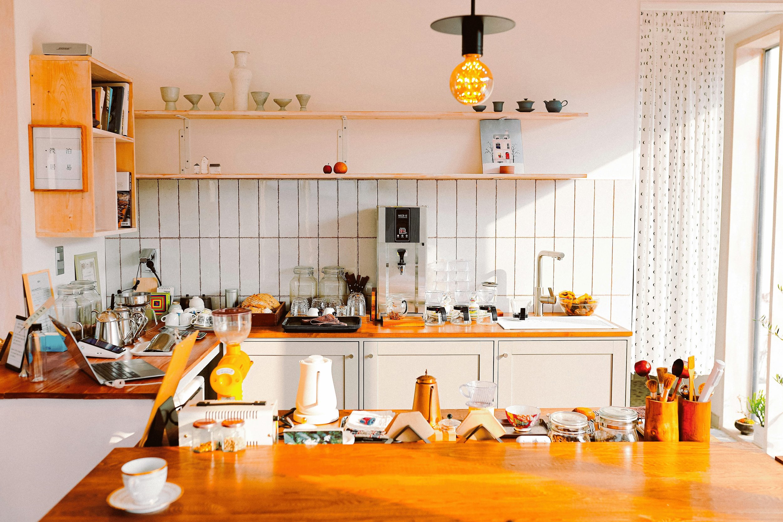 A kitchen scene with a wooden countertop cluttered with cups, jars, and kitchen tools, a white tiled backsplash, and open shelving with decorative items and a hanging orange light. Lighter Home decluttering services available in Gainesville, Florida.