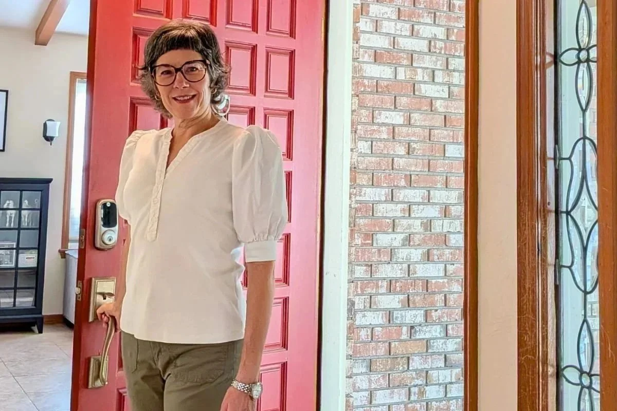 Lighter Home founder Colleen Raccioppi walking through a client's front door. She has short curly hair, wearing glasses, a white blouse with puffed sleeves, khaki pants, and a watch, standing at the open front door of a house.