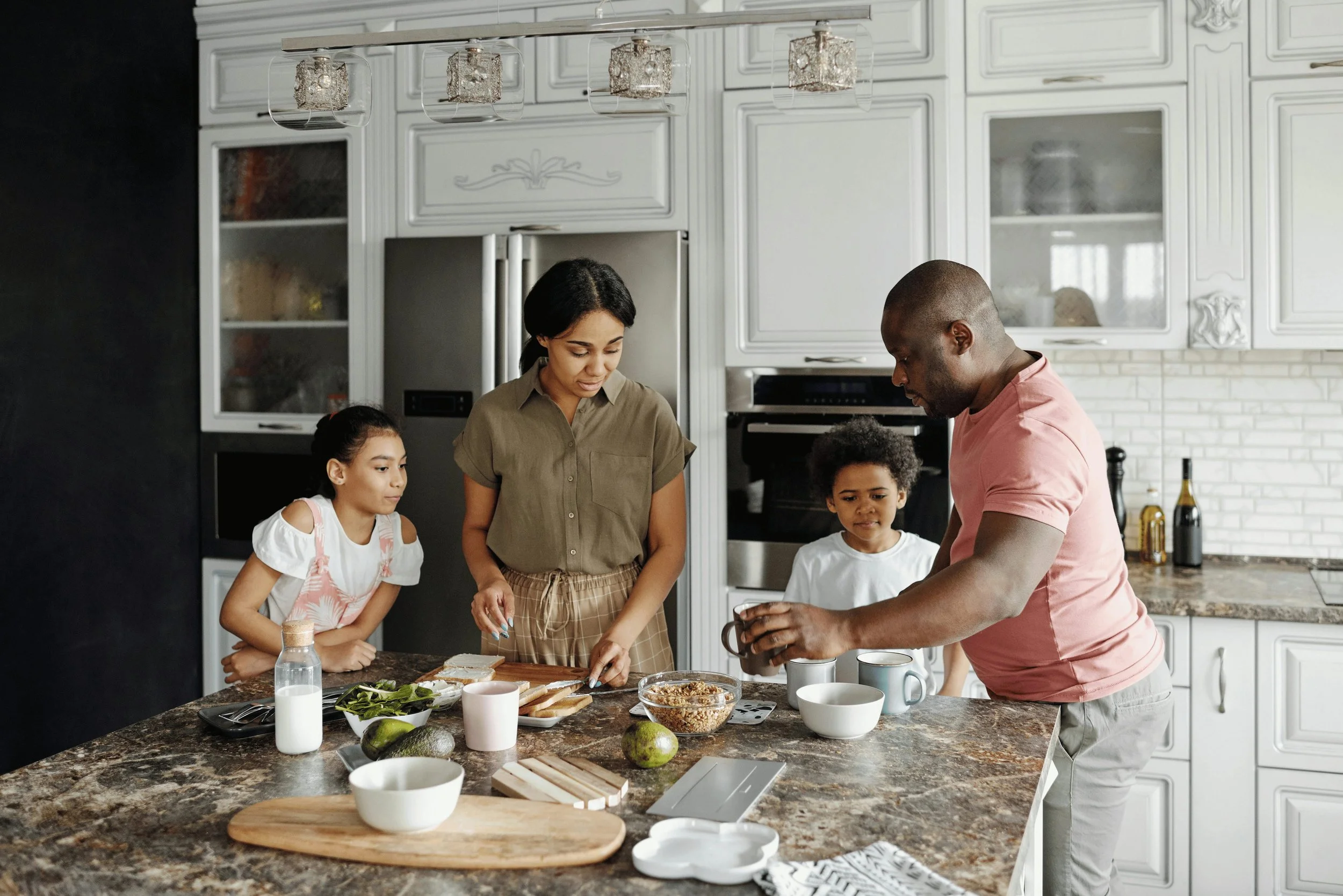 A family of four preparing breakfast in a modern kitchen. The mother and two children are gathered around the counter, and the father is pouring a drink. Lighter Home is for busy families where the time and energy to run the house is in short supply.
