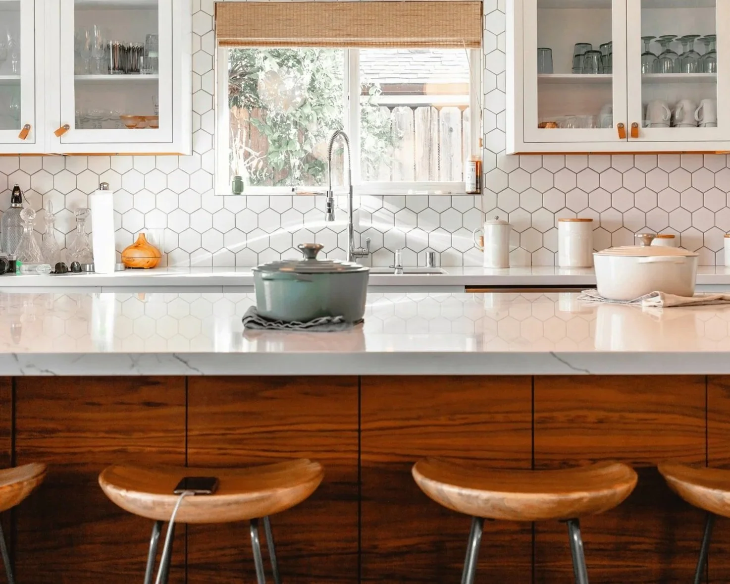 Lighter Home kitchen reset showing a clean kitchen with white hexagonal tile backsplash, wooden cabinets, marble countertop island with stools, and open shelves with dishes and glassware; window above sink with greenery outside.