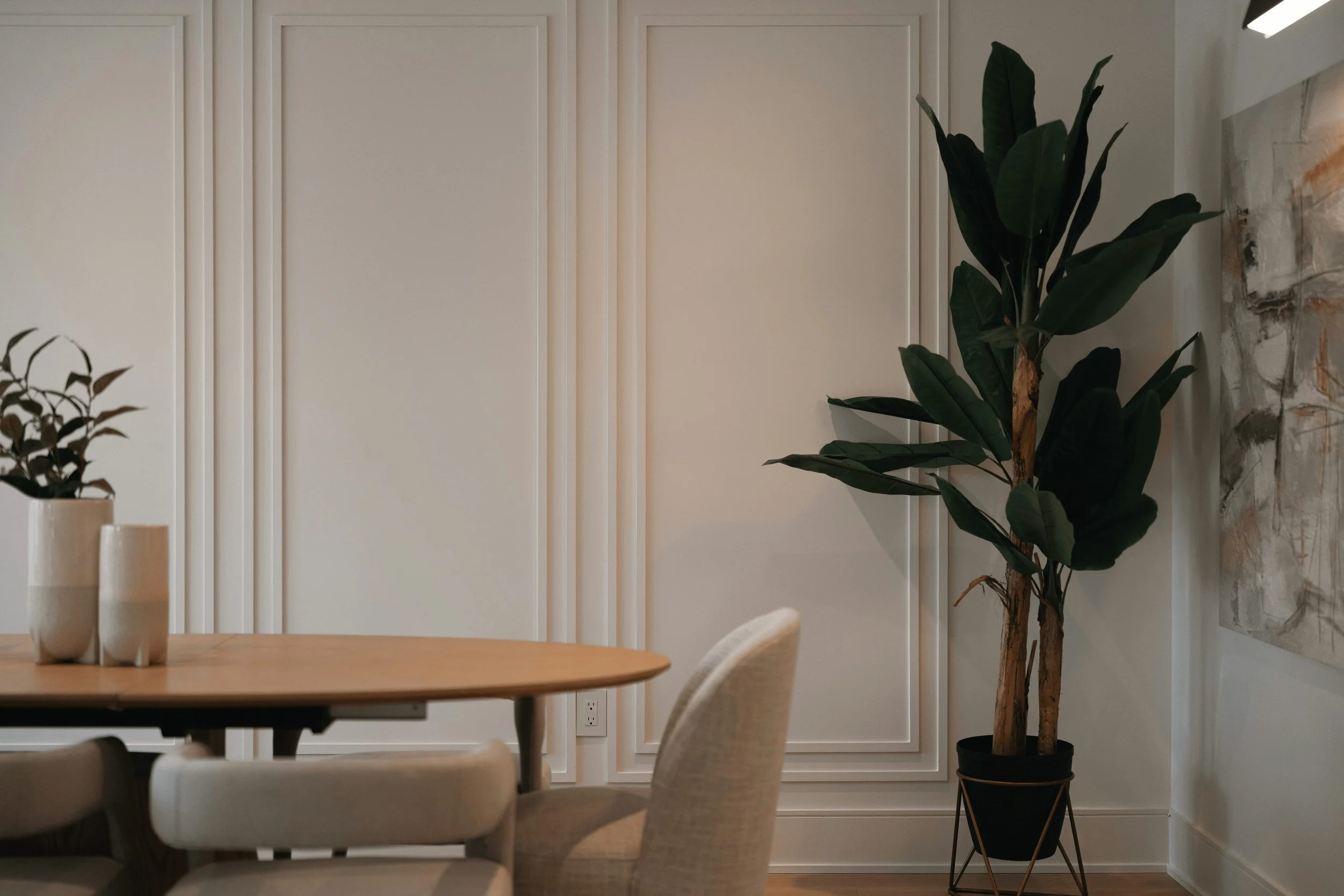 Modern dining room with a large potted plant, beige upholstered chairs, a wooden table, and decorative wall panels.