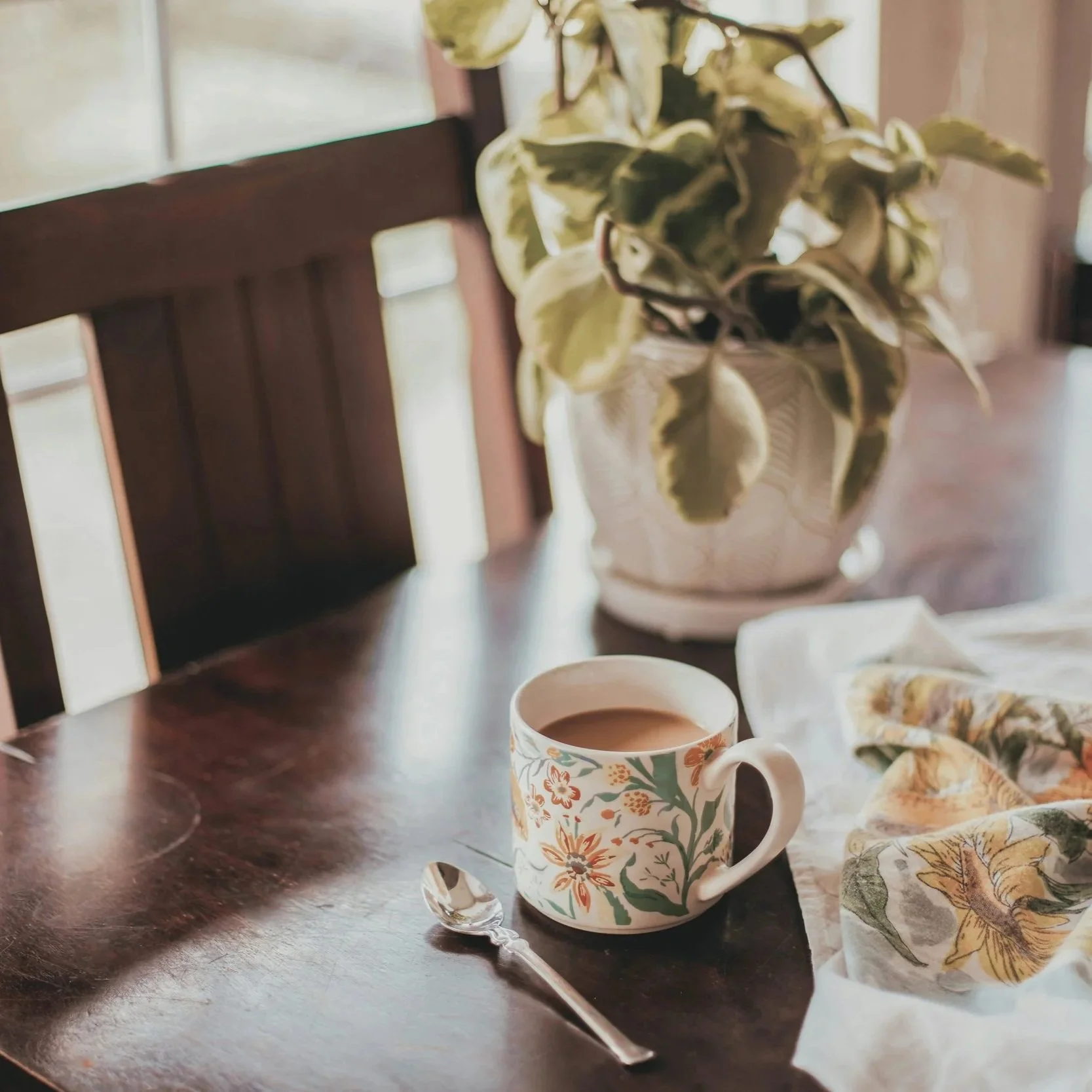 A table with a floral ceramic mug filled with a hot beverage, a silver teaspoon, a cloth with a leaf pattern, a plate with cookies, and a potted plant. Unhurried morning coffee at home — time returned by Lighter Home household management service.
